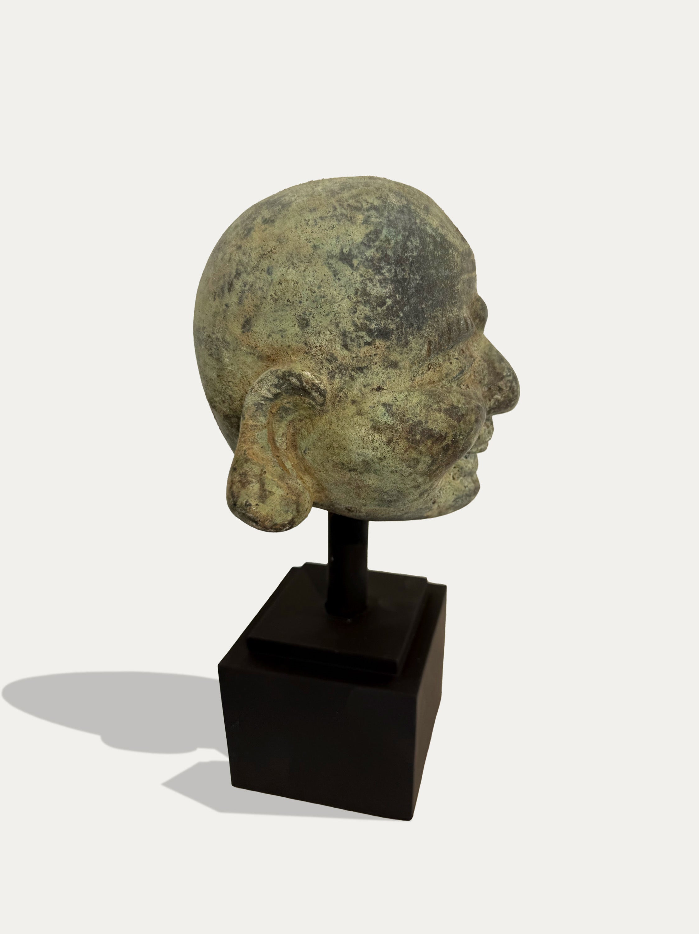 An ancient-style bronze or stone head sculpture shown in profile on a black display stand against a light gray background.