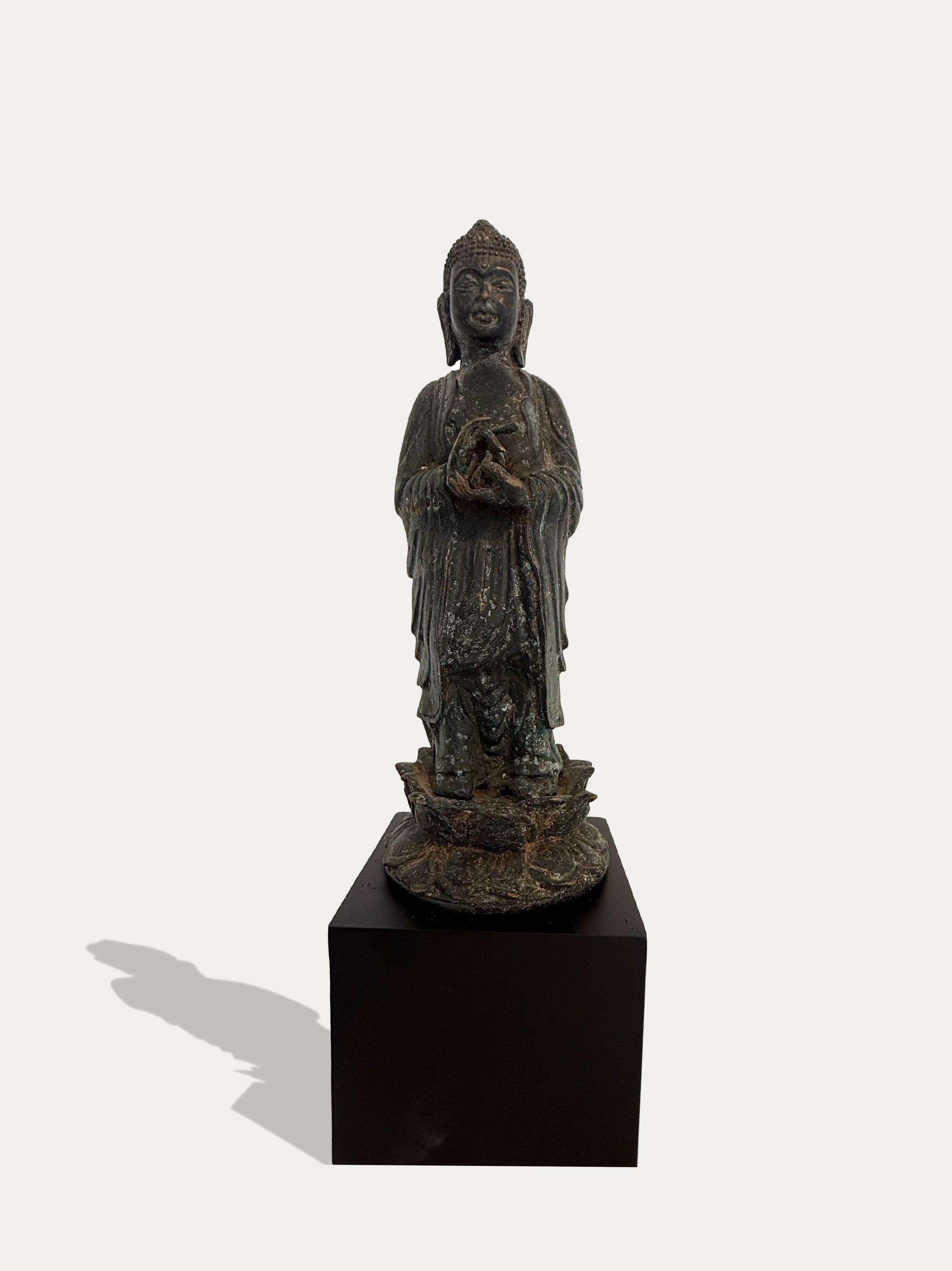 A dark bronze standing Buddha statue with a weathered patina, displayed on a black rectangular pedestal against a light gray background.