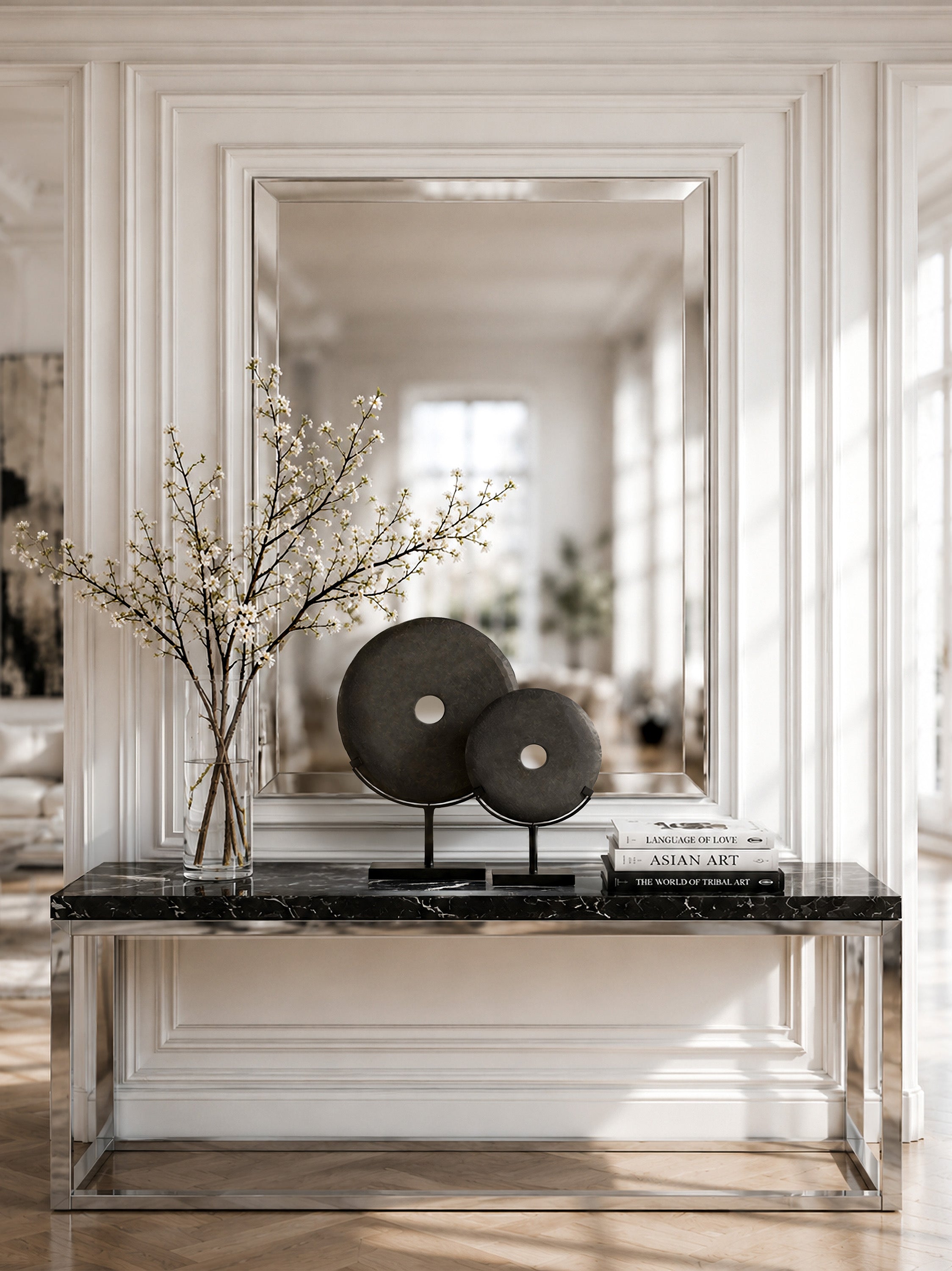 A styled interior scene with a large rectangular mirror mounted on a white paneled wall above a console table. On the table sits a black marble surface with two round dark decorative objects on black stands, a stack of books, and a clear glass vase holding delicate white flowering branches.