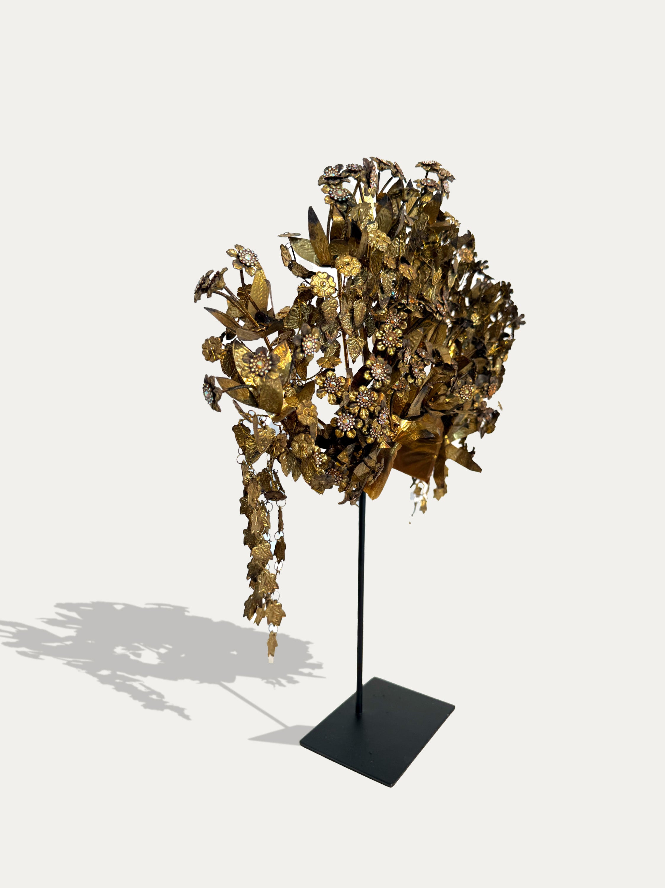 A decorative gold-toned metal tree sculpture with hanging leafy branches, mounted on a slim black stand with a square base, photographed against a plain light gray background.
