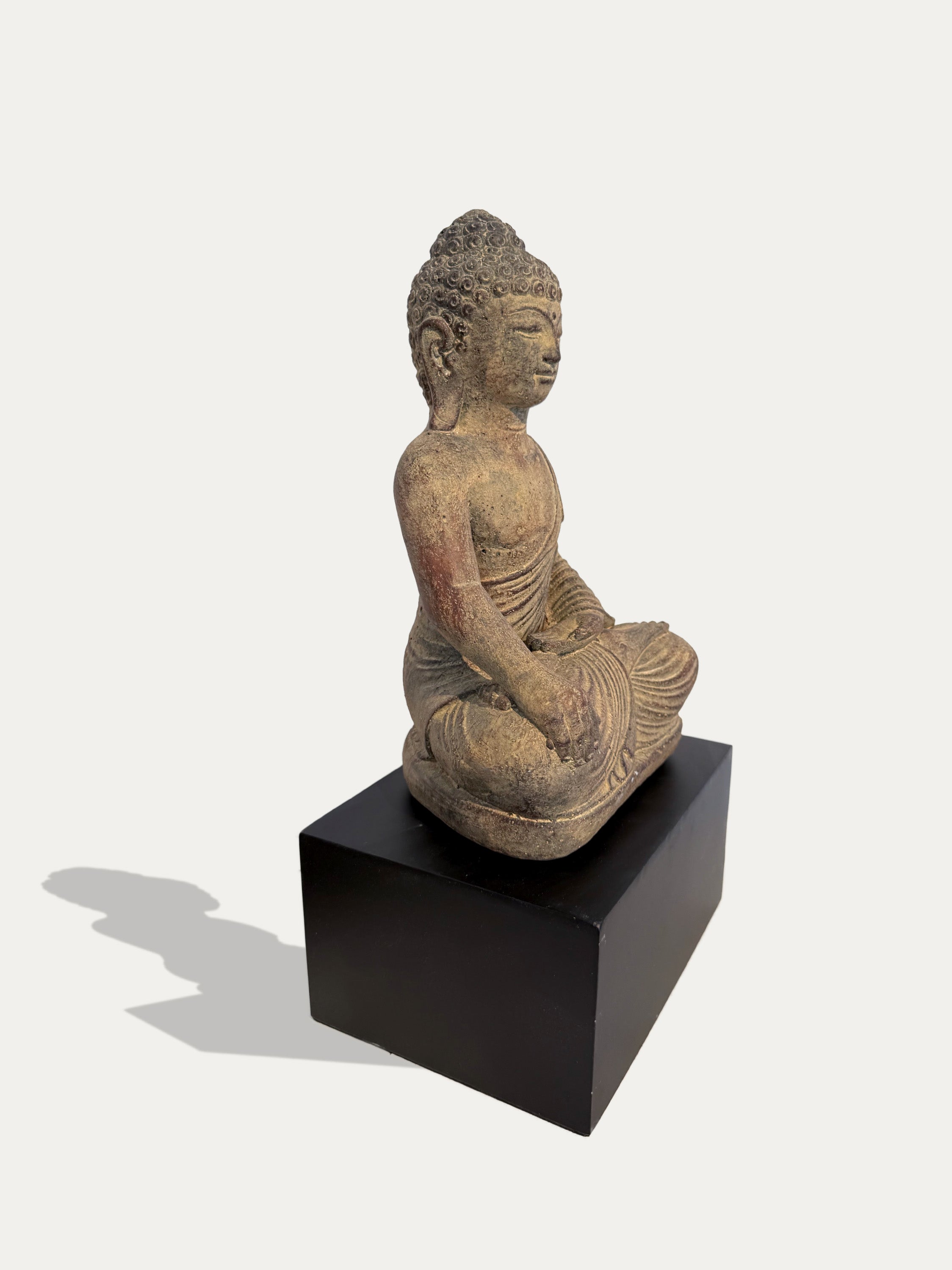 A seated Buddha statue in a weathered tan and brown stone-like finish, displayed on a black rectangular pedestal against a light gray background.