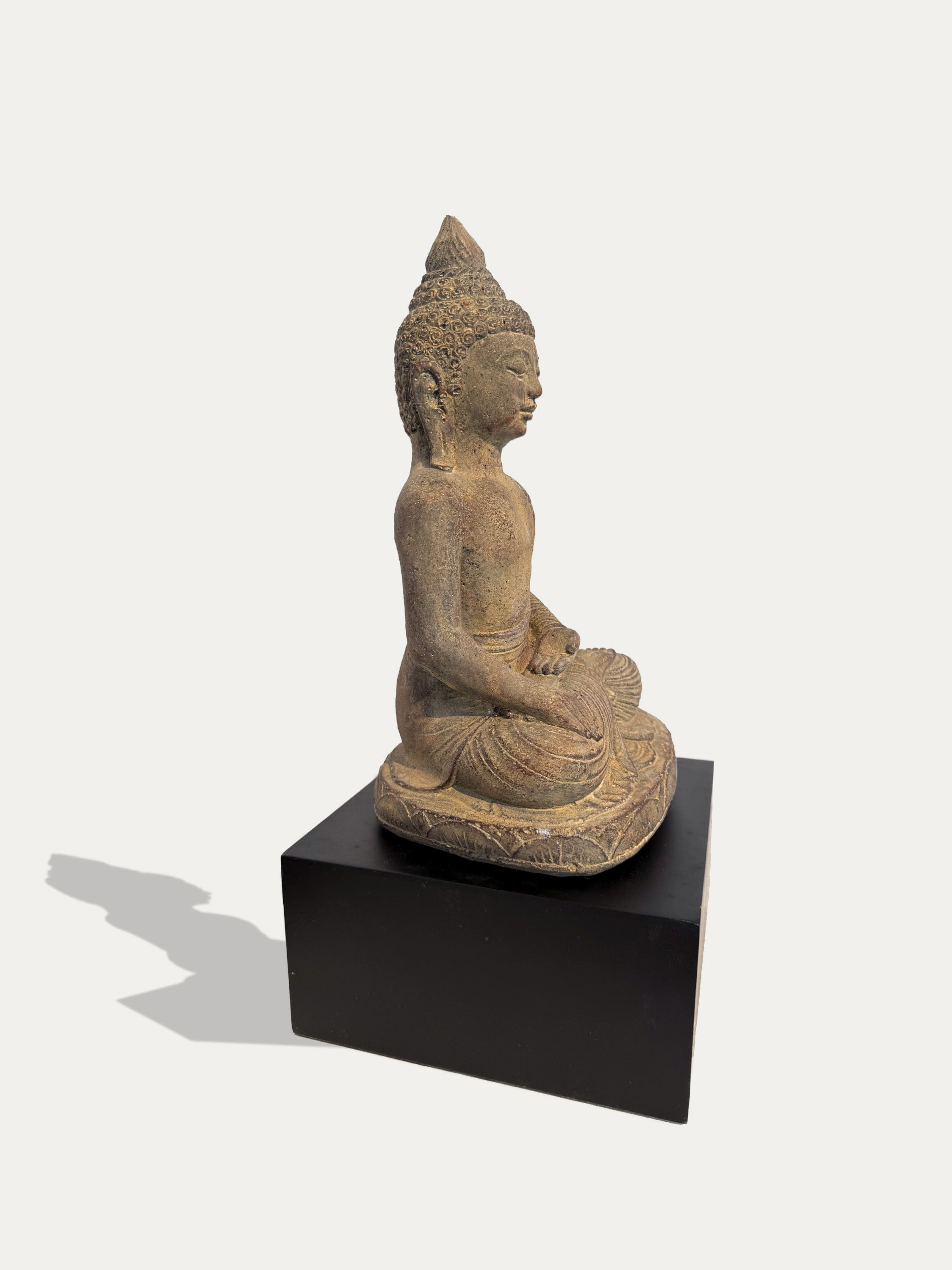An antique-style seated Buddha statue in a weathered beige stone or concrete finish, displayed on a solid black rectangular pedestal against a light gray background.