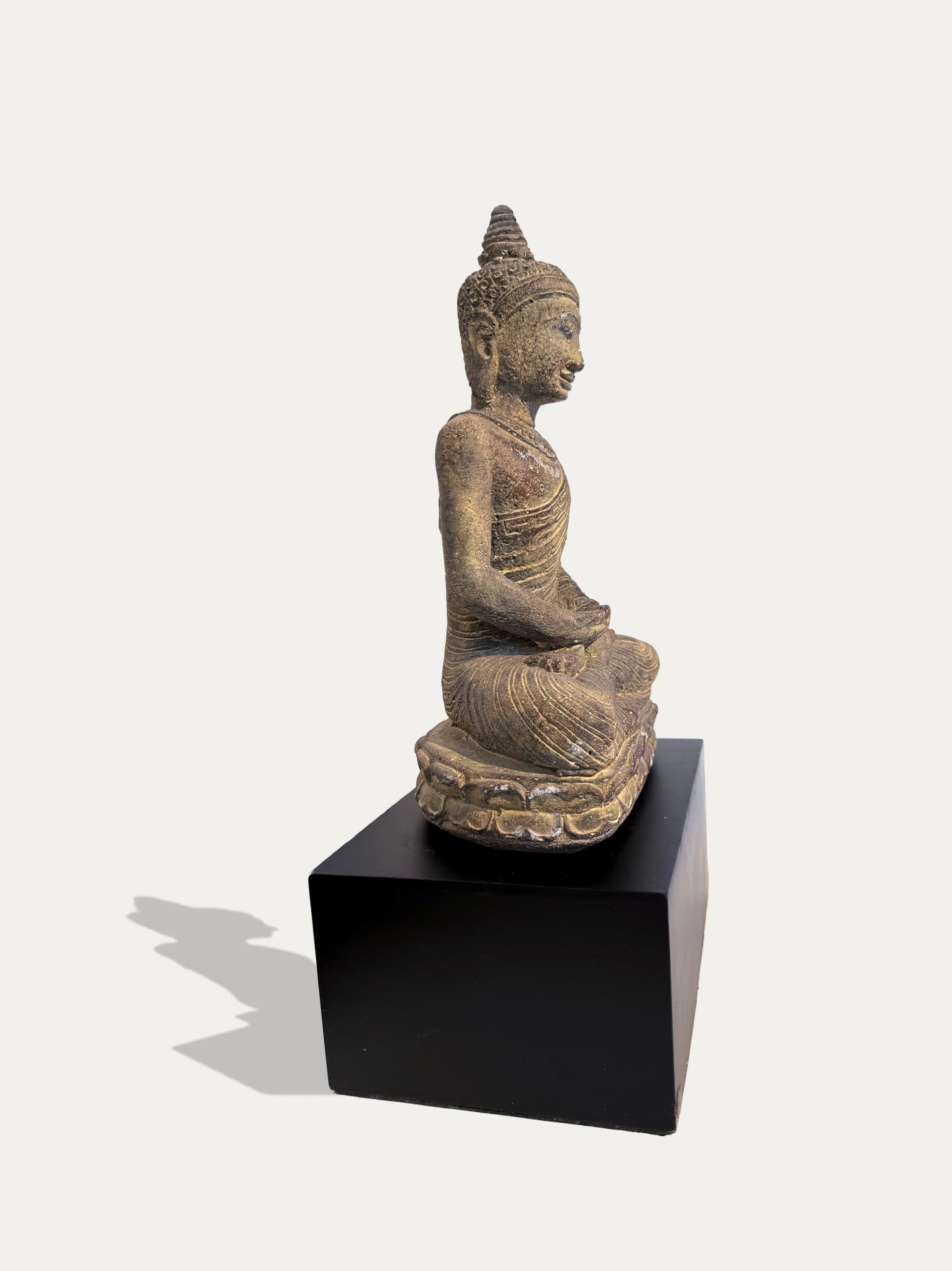 A seated Buddha statue in a meditative pose, shown in profile on a black display pedestal against a plain light background.