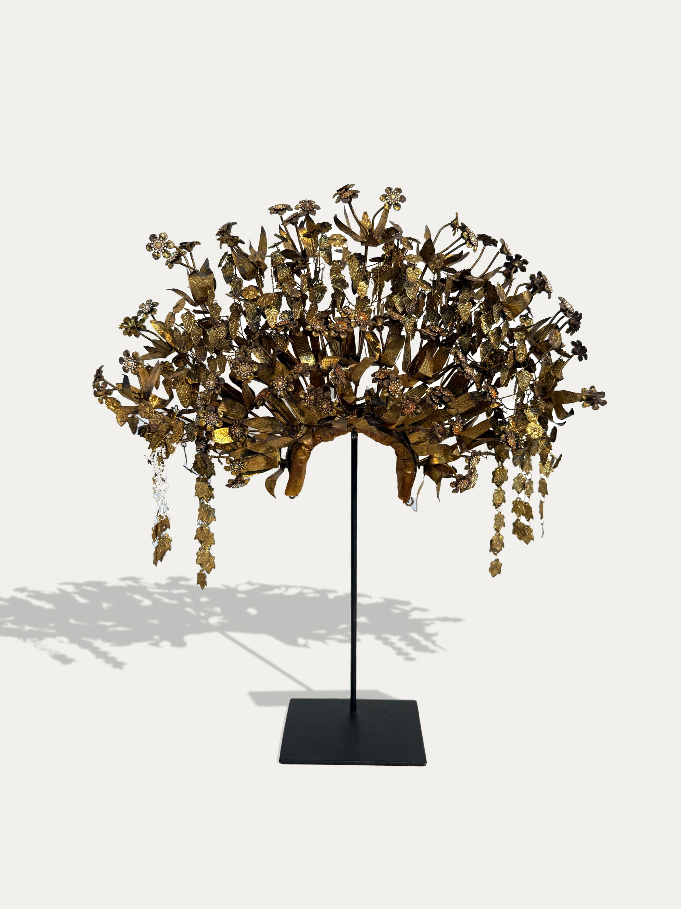 A gold-toned floral crown or headdress displayed on a black stand against a light gray background. The piece features an intricate arrangement of metallic gold leaves and hanging flower-like elements, with a black rectangular base.