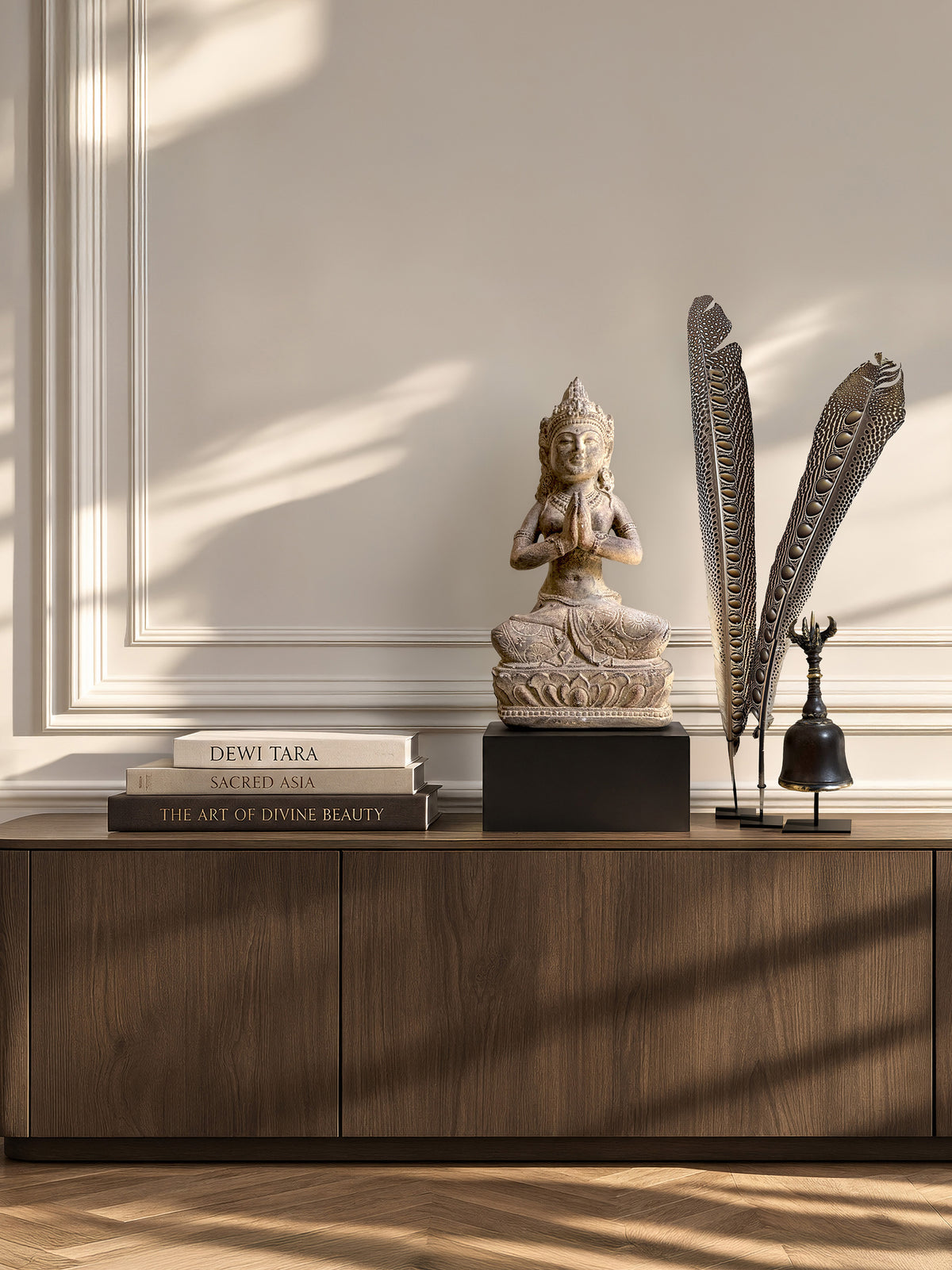 Conjunto Divine Grace con estatua de Dewi Tara, dos plumas de pavo real y una campana Ghanta sobre una consola de madera iluminada por el sol