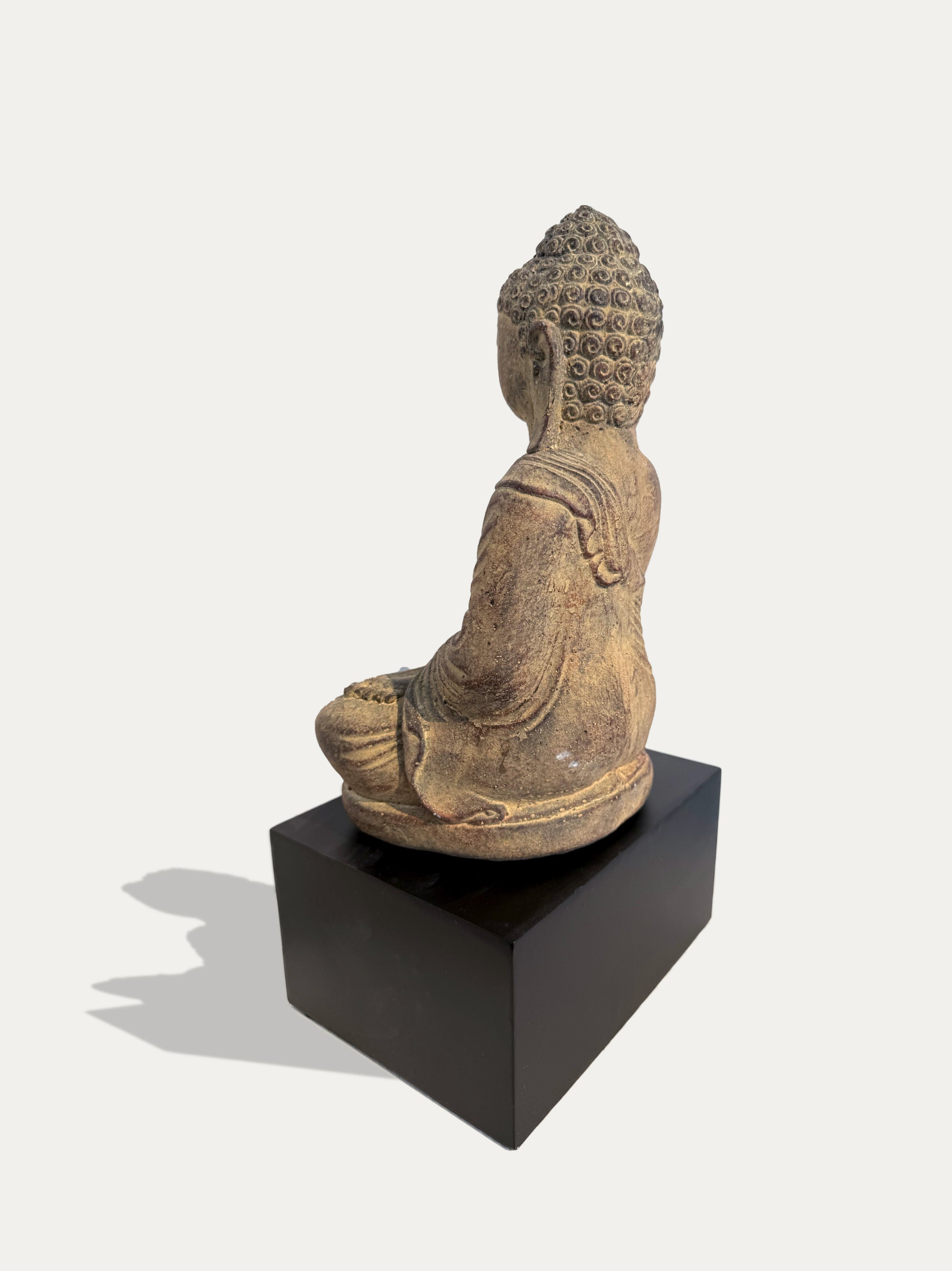 A seated Buddha statue in a weathered tan and brown stone-like finish, shown from the back on a black rectangular pedestal against a light gray studio background.
