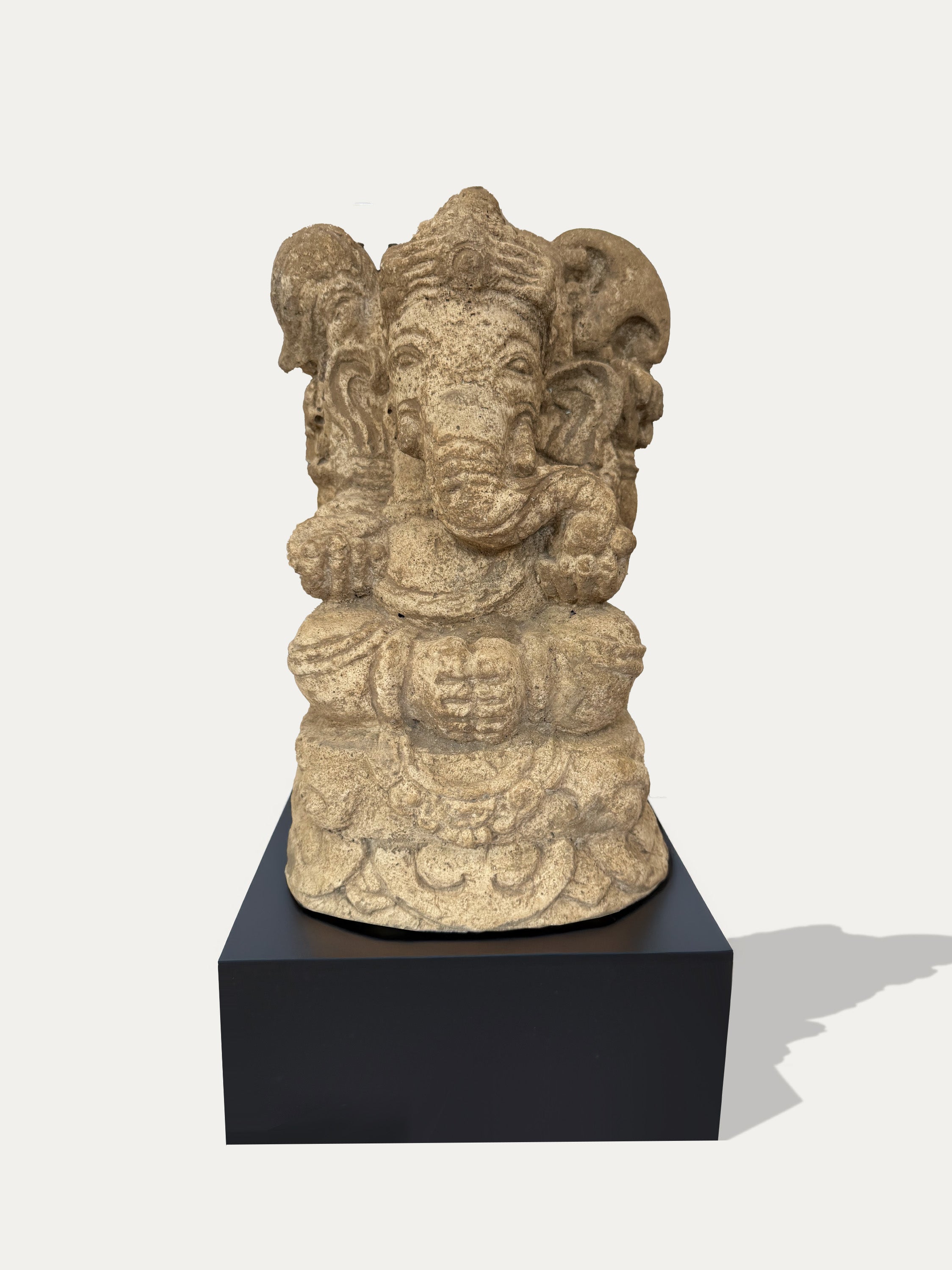 A carved stone Ganesha statue in a light beige/tan finish, displayed on a black rectangular pedestal against a light gray background.