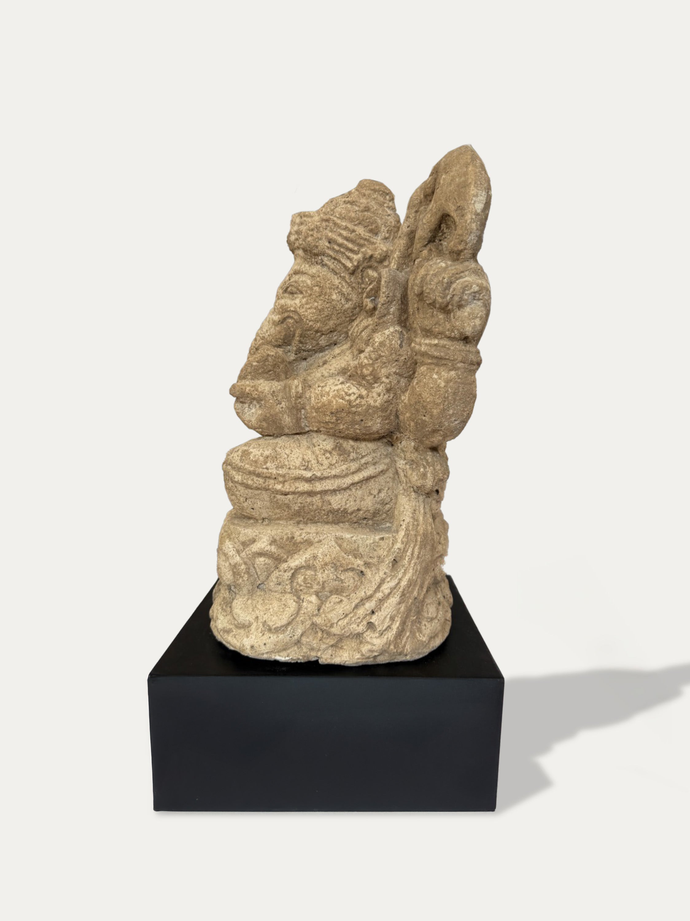 A carved stone statue of a seated figure with a tall headdress and curved back, displayed on a black rectangular pedestal against a light gray background.