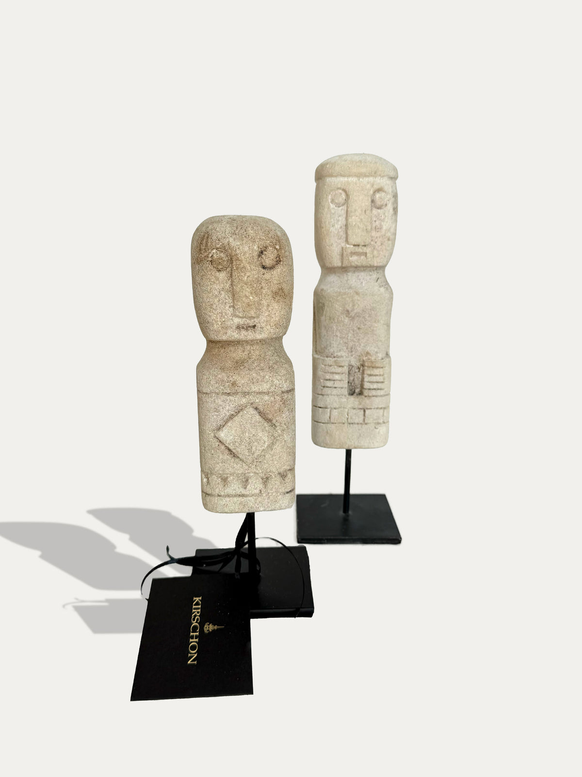 Front view of two hand-carved tribal limestone statues from Timor, Indonesia, on black metal stands.