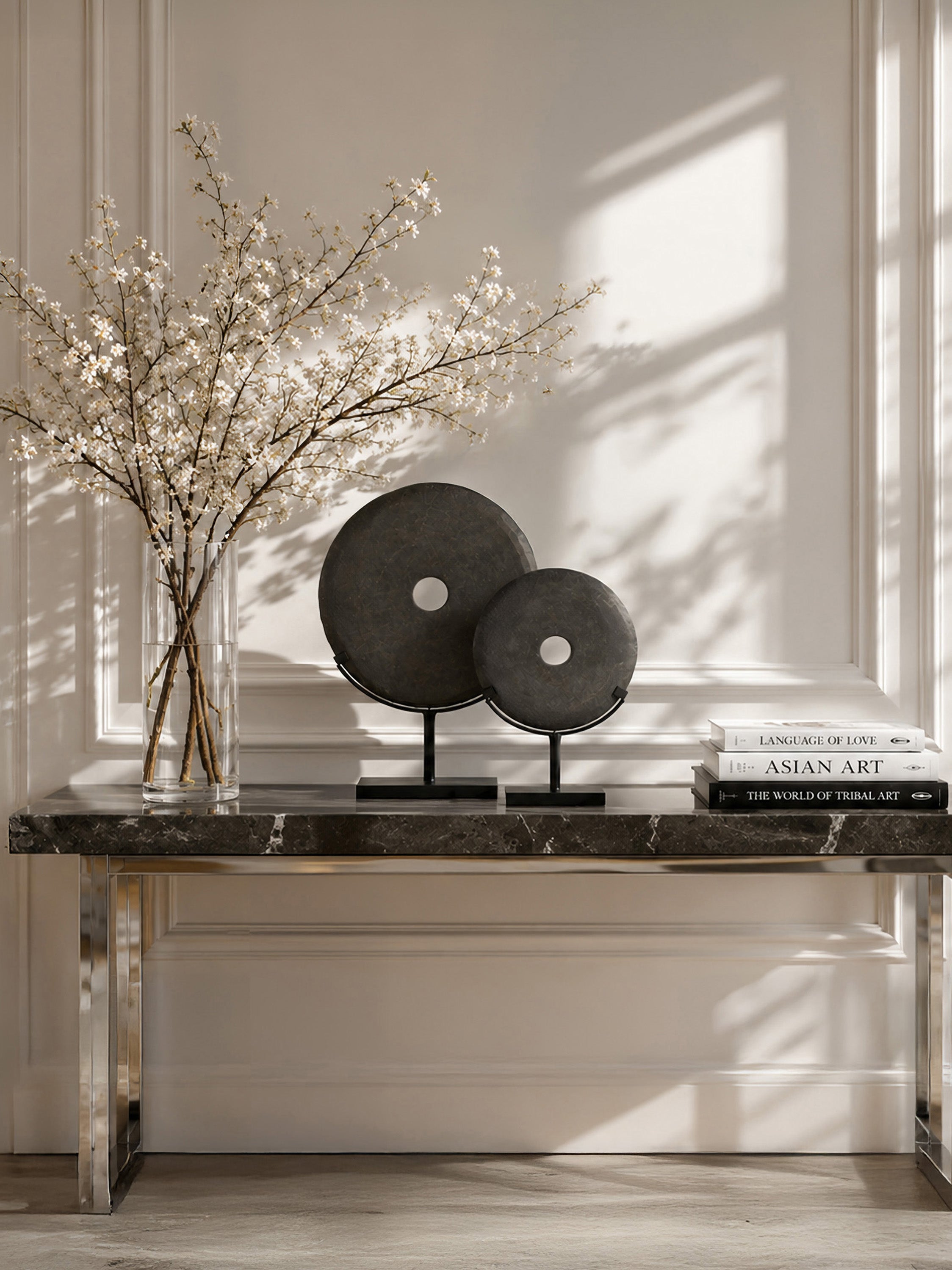 Two round black stone sculptures on black stands displayed on a marble console table in a bright room with sunlight and shadows, next to a glass vase with dried white blossoms and a stack of books.