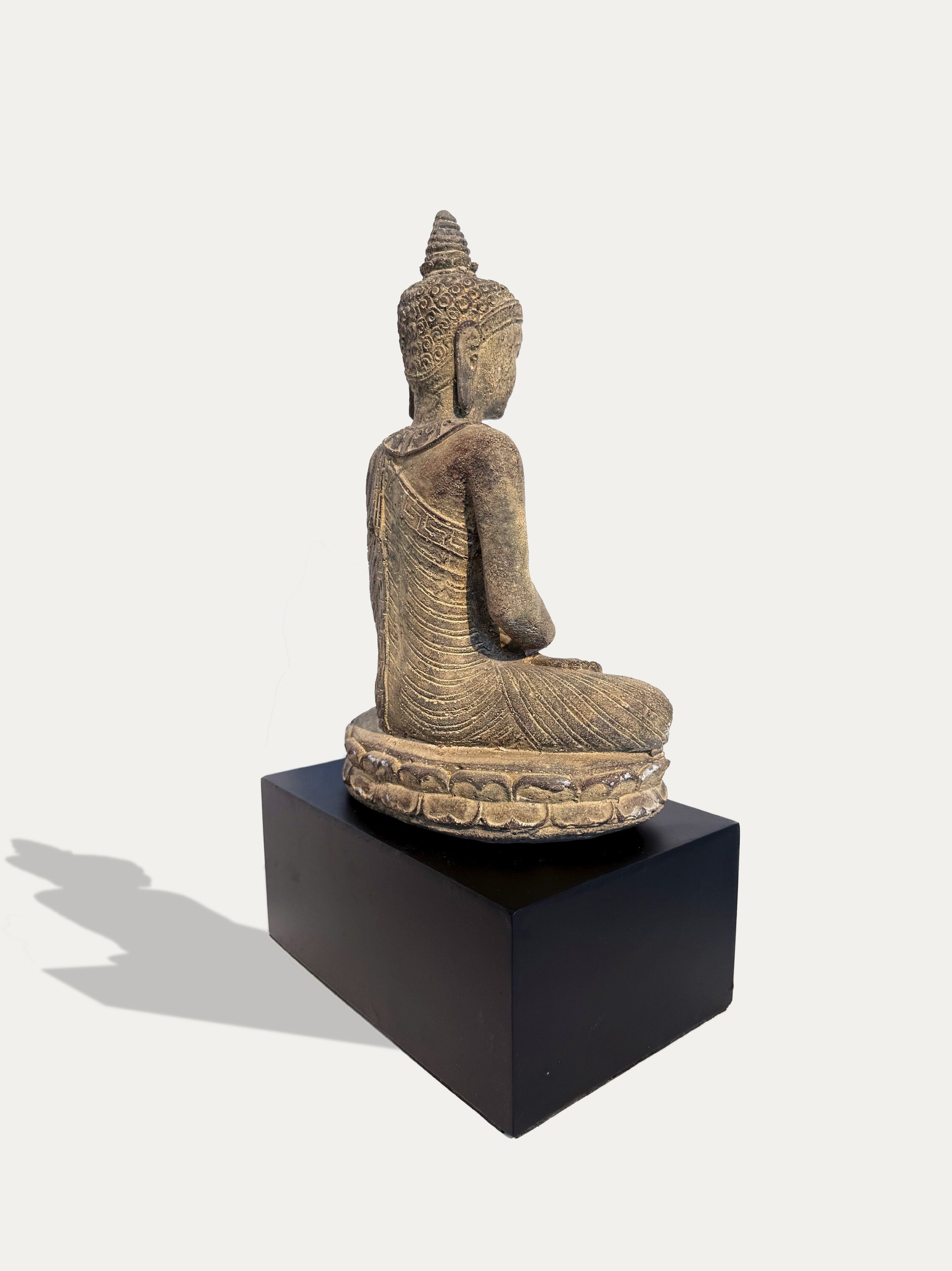 A seated Buddha statue in a meditative pose, shown from the back on a black pedestal against a light gray background.