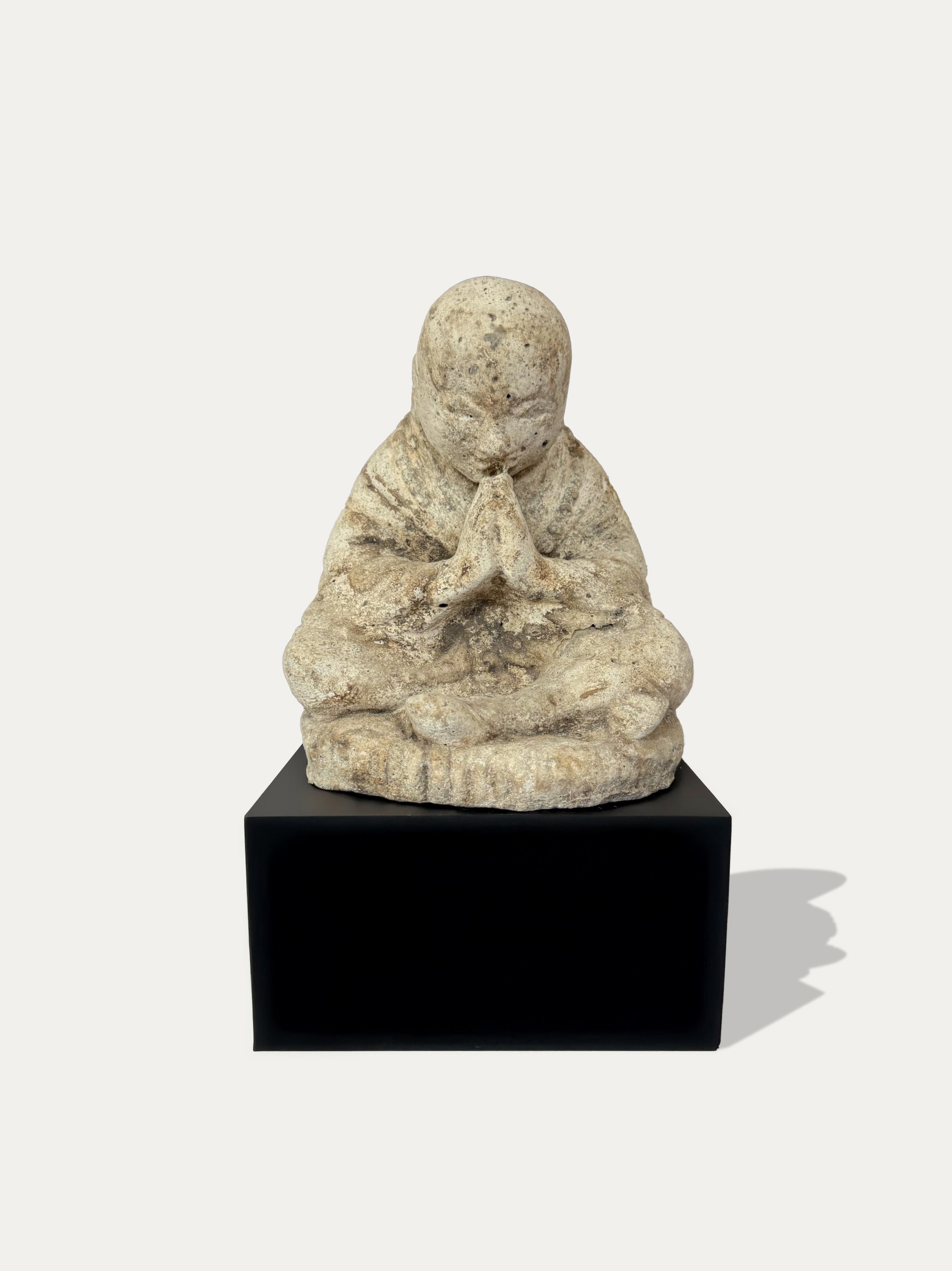 A small beige stone figurine of a seated, prayer-holding child, displayed on a black rectangular pedestal against a light gray background.