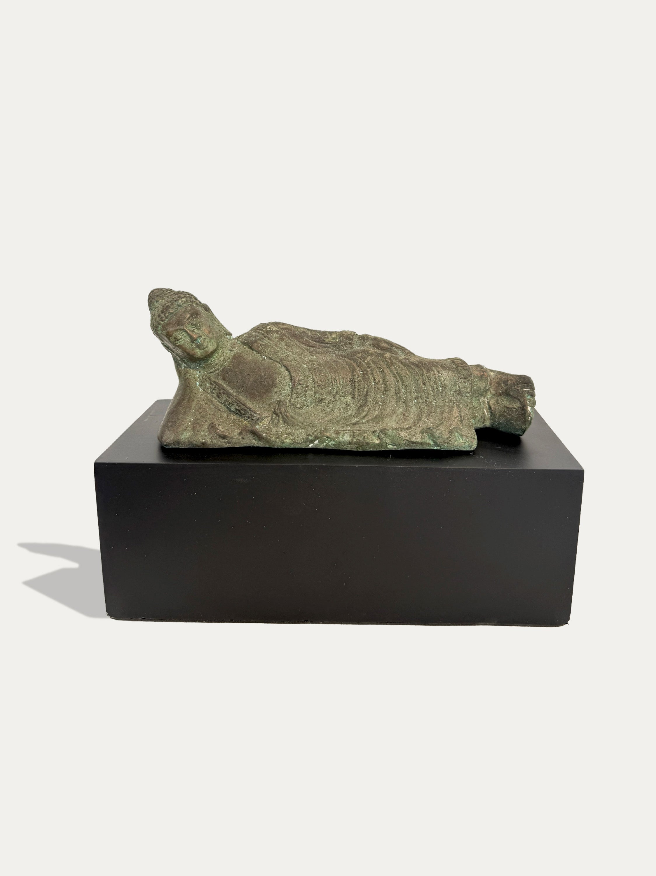 A small reclining Buddha statue with a weathered greenish-bronze patina, displayed on a dark rectangular pedestal against a light gray background.