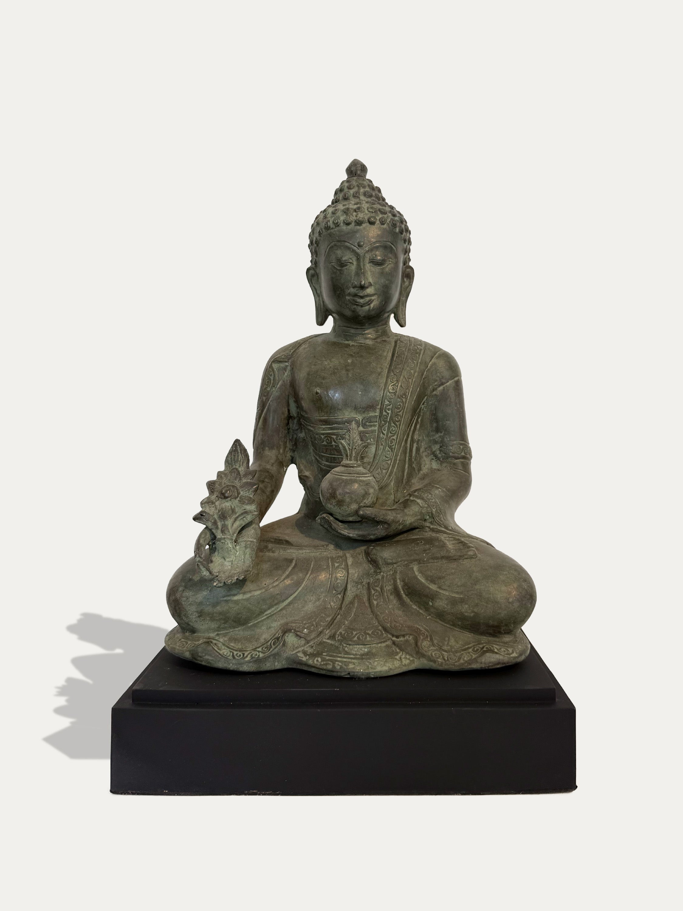 A seated Buddha statue in an aged bronze finish, positioned on a solid black rectangular base against a light off-white background.