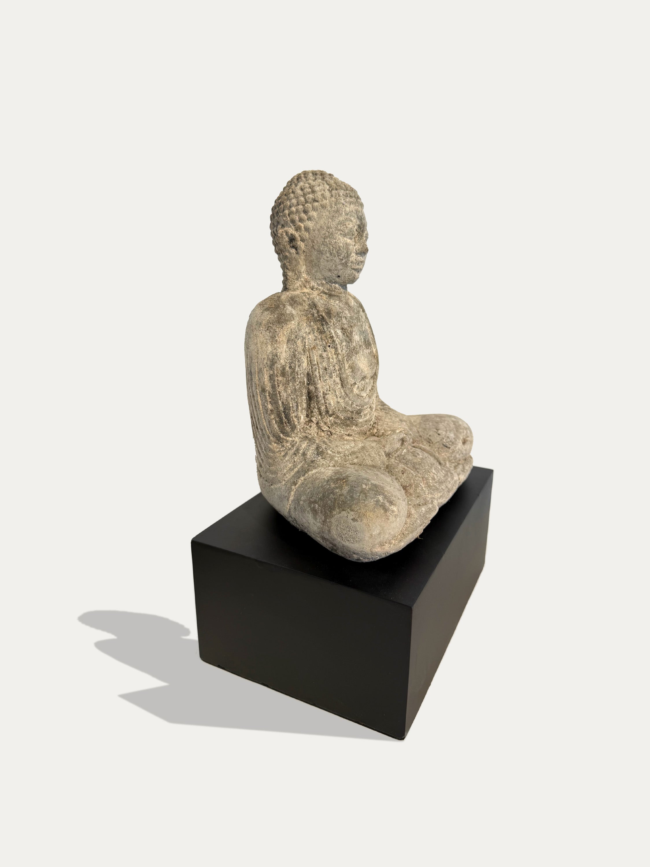 A seated Buddha statue in a light beige stone-like finish, displayed on a black rectangular pedestal against a light gray background.