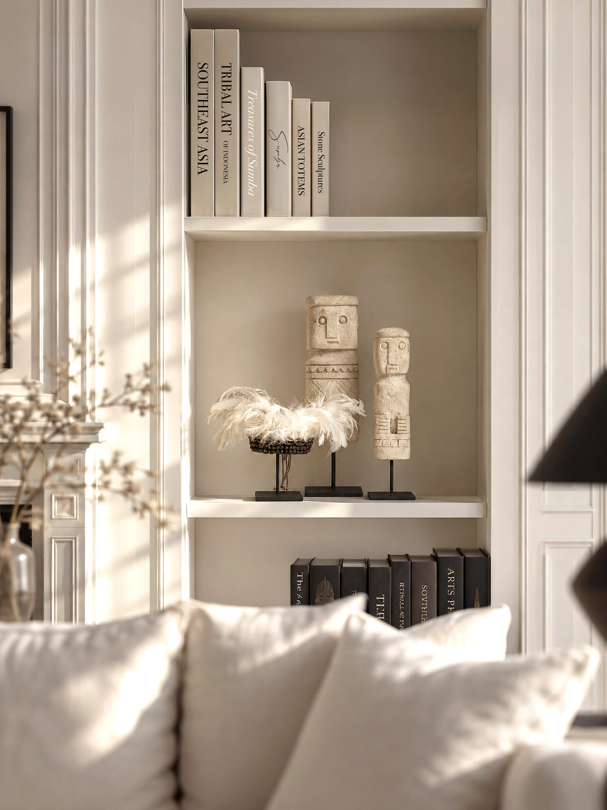 Two Timor stone totems and a Papua feather-and-shell bracelet displayed on a white bookshelf behind a sofa