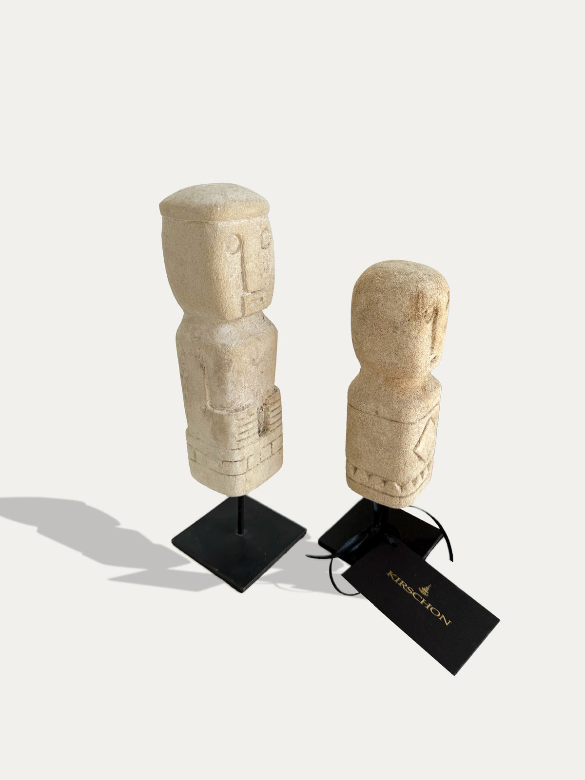 Angled view of two tribal limestone statues from Timor, showing carved faces and geometric patterns on black metal stands.