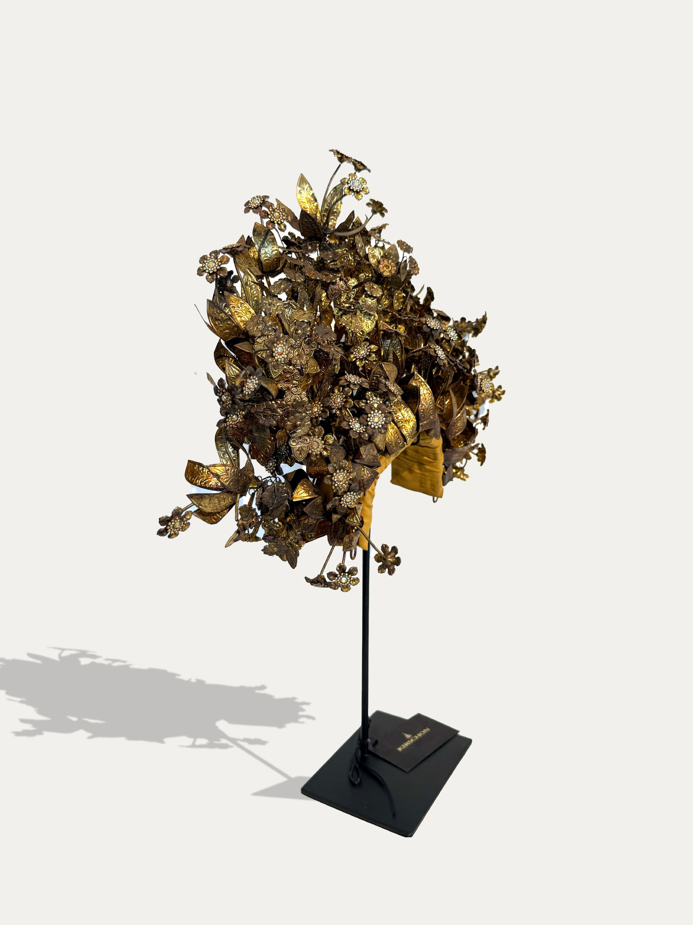 A gold-toned floral headpiece with metallic leaves and ribbon accents, displayed on a black stand against a light gray background.