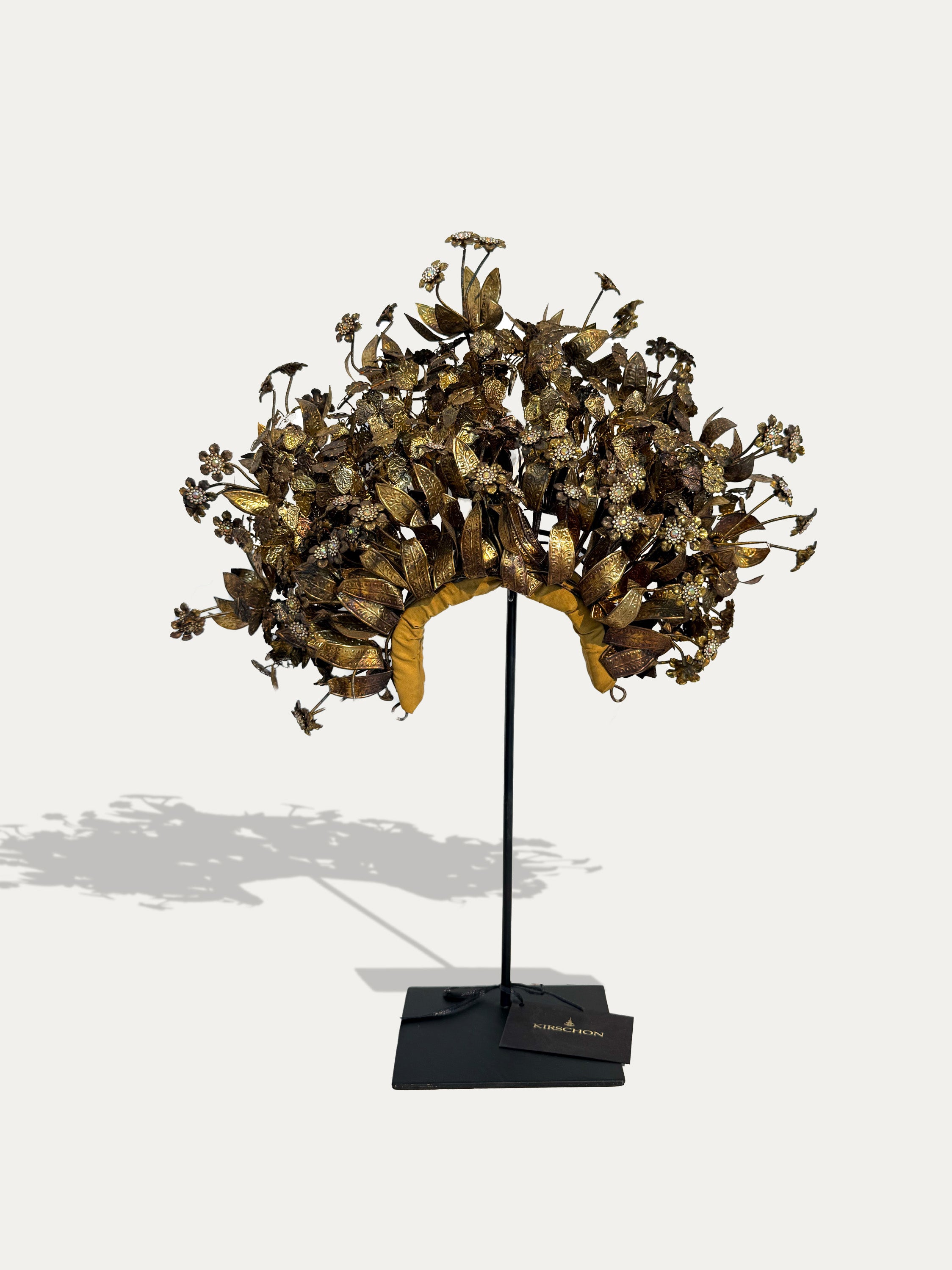 An ornate vintage-style metal floral crown or headdress displayed on a black stand against a light gray background. The piece features intricate gold-toned leaf and flower motifs with a curved yellow-gold base, mounted on a black display stand with a small plaque at the base.