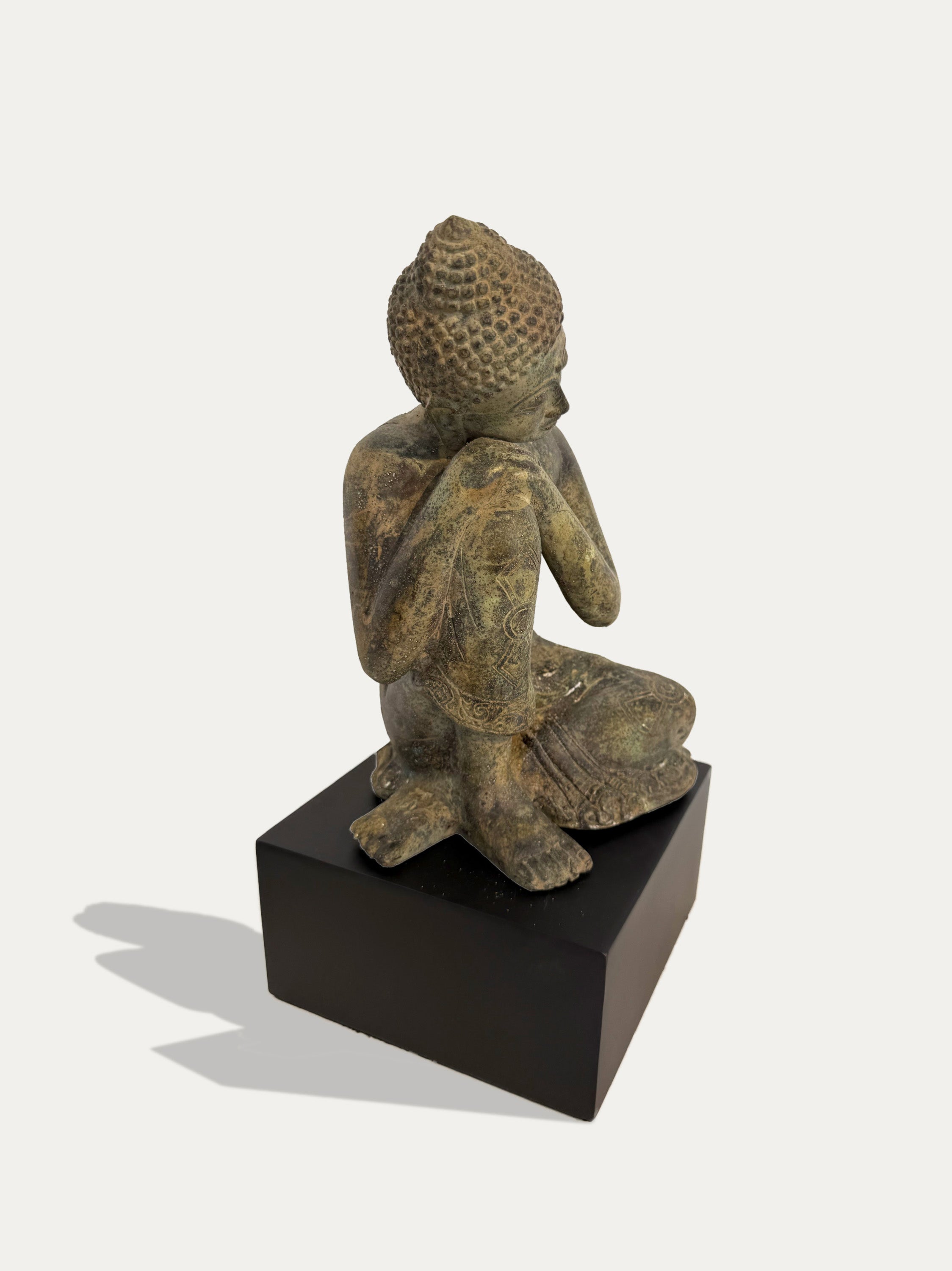 A small seated Buddha statue with a textured, aged metallic finish, mounted on a rectangular black base against a light gray background.