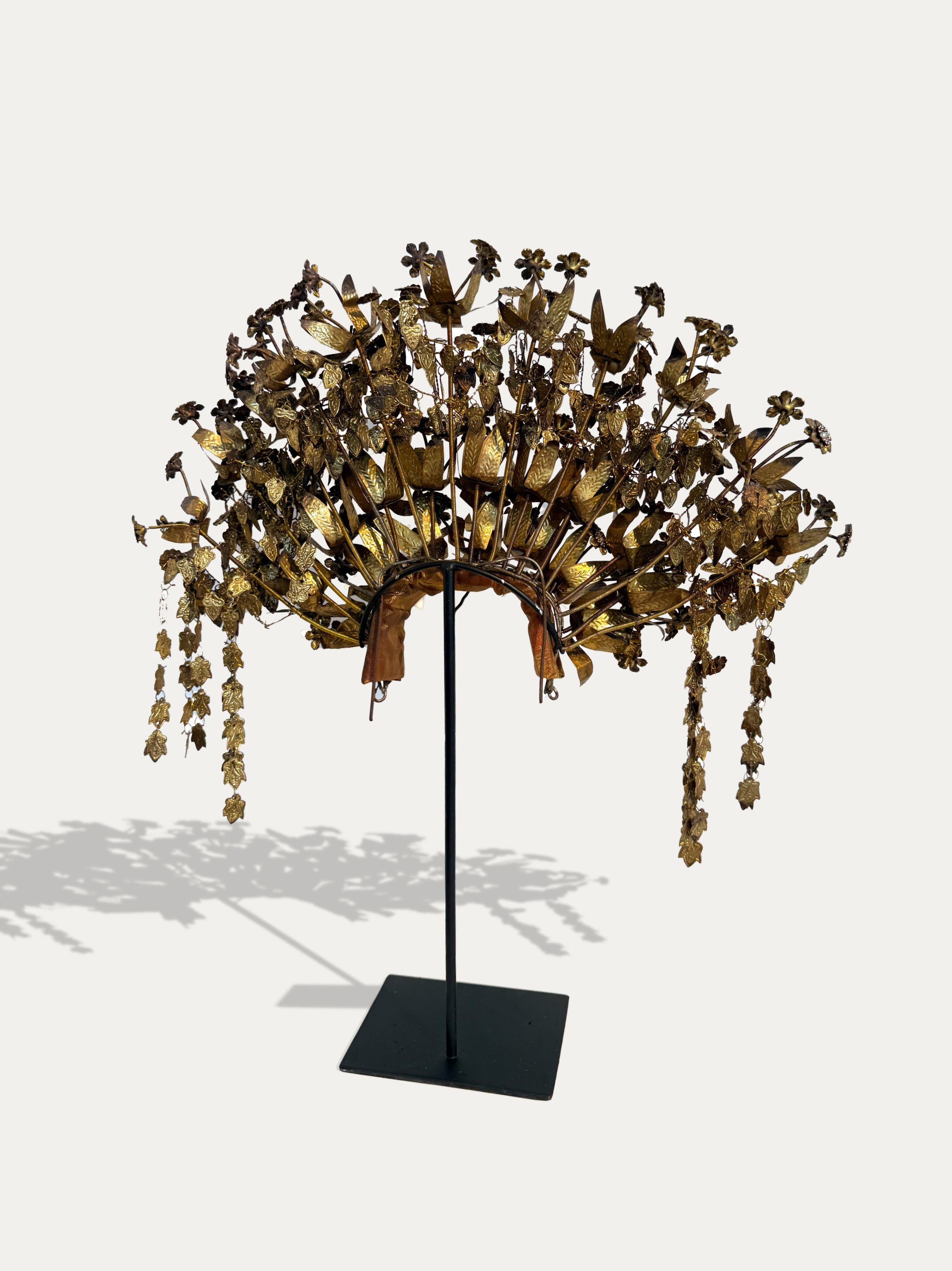 A large ornate gold-toned floral crown or headdress displayed on a black stand against a light gray background. The piece features many layered flower-like elements and hanging dangling chains.