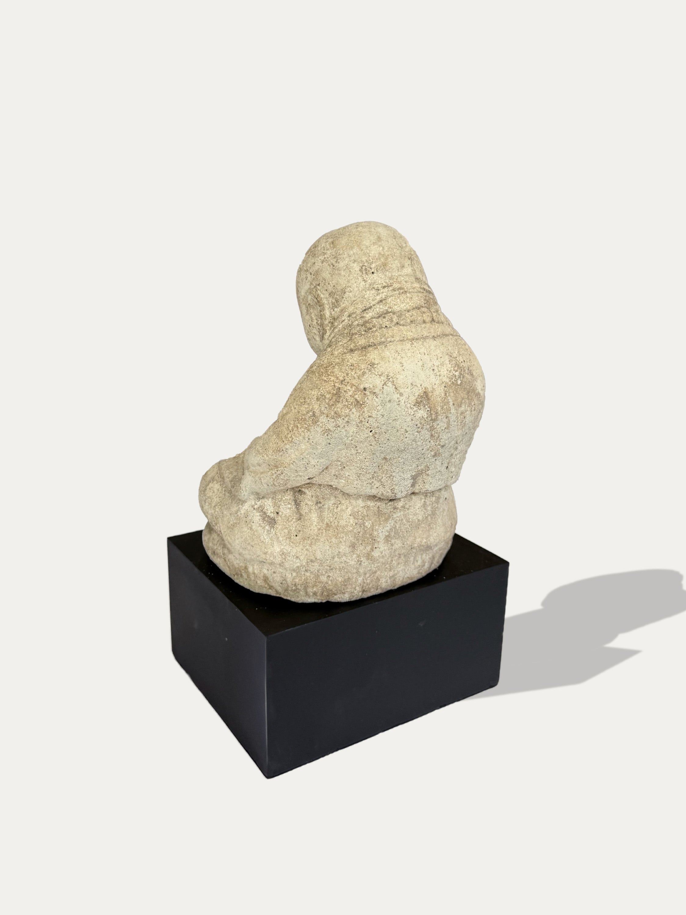 A small beige stone figurine of a seated robed figure, shown from the back, mounted on a black rectangular pedestal against a light gray background.