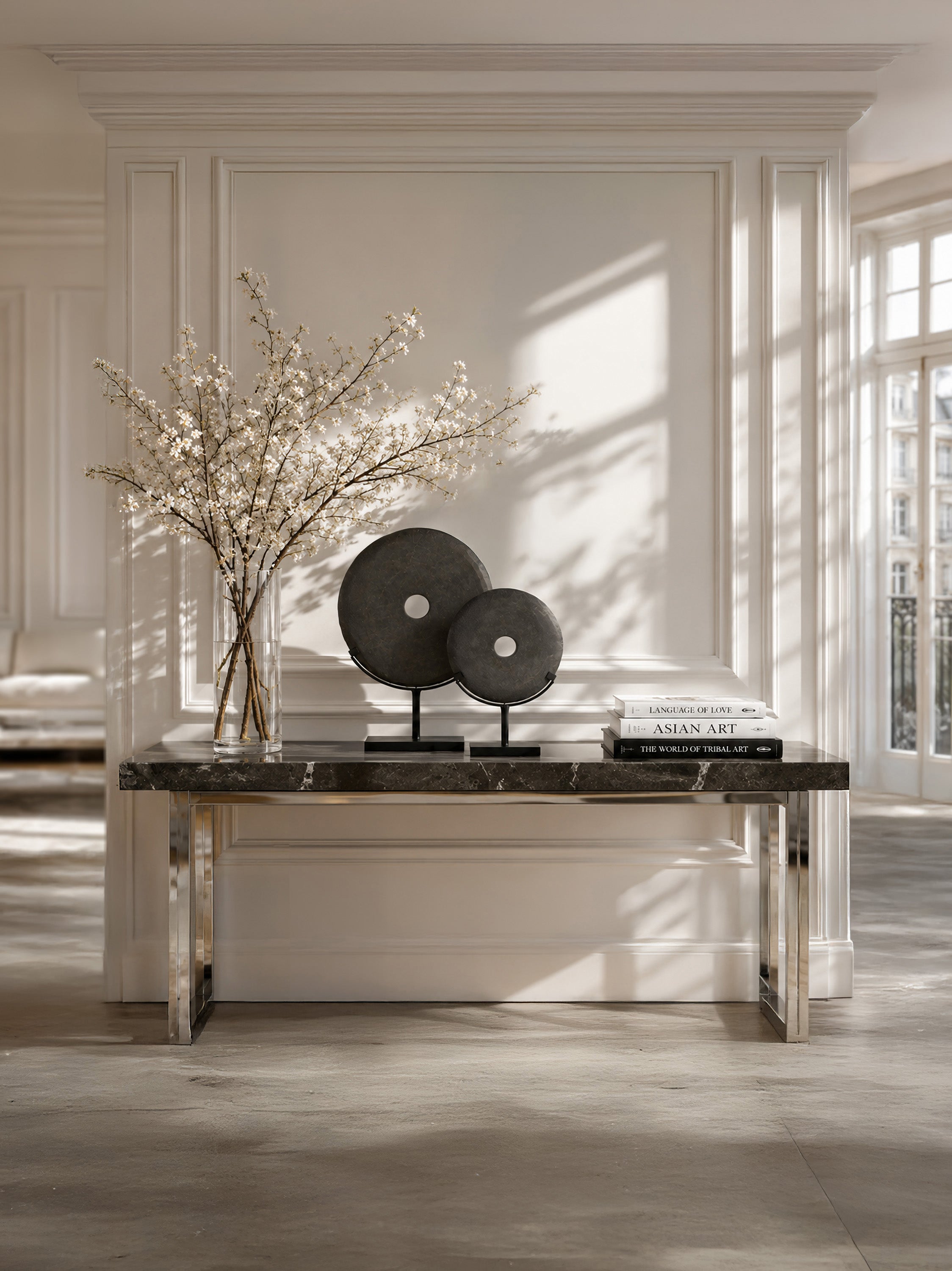 A modern console table with a dark marble top and polished metallic legs, styled in a bright, elegant room with sunlight streaming through windows. Decorative elements include a large vase with white blossoms and two round black art pieces.