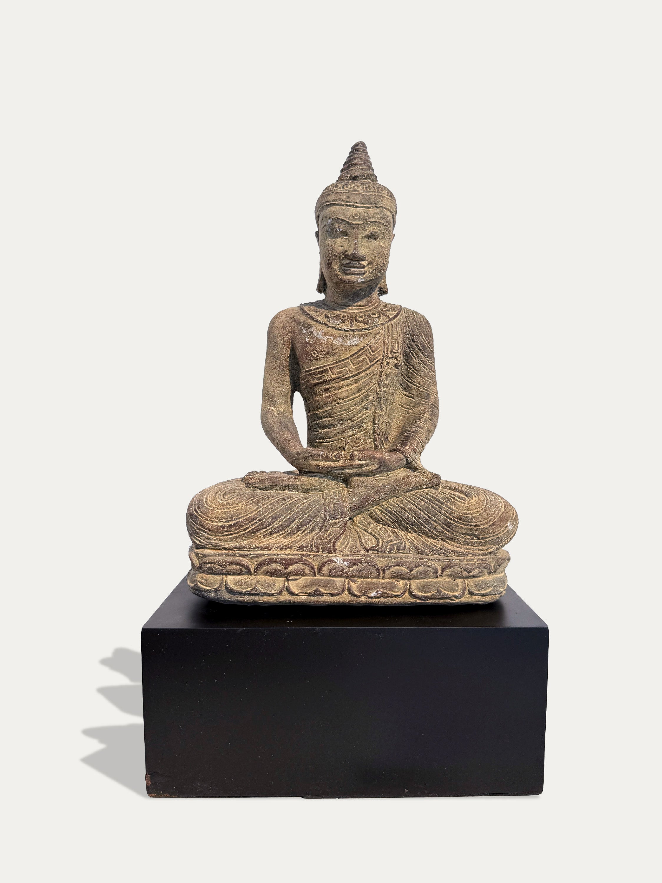 An antique-style seated Buddha statue with a weathered brown and beige patina, displayed on a solid black rectangular pedestal against a light off-white background.