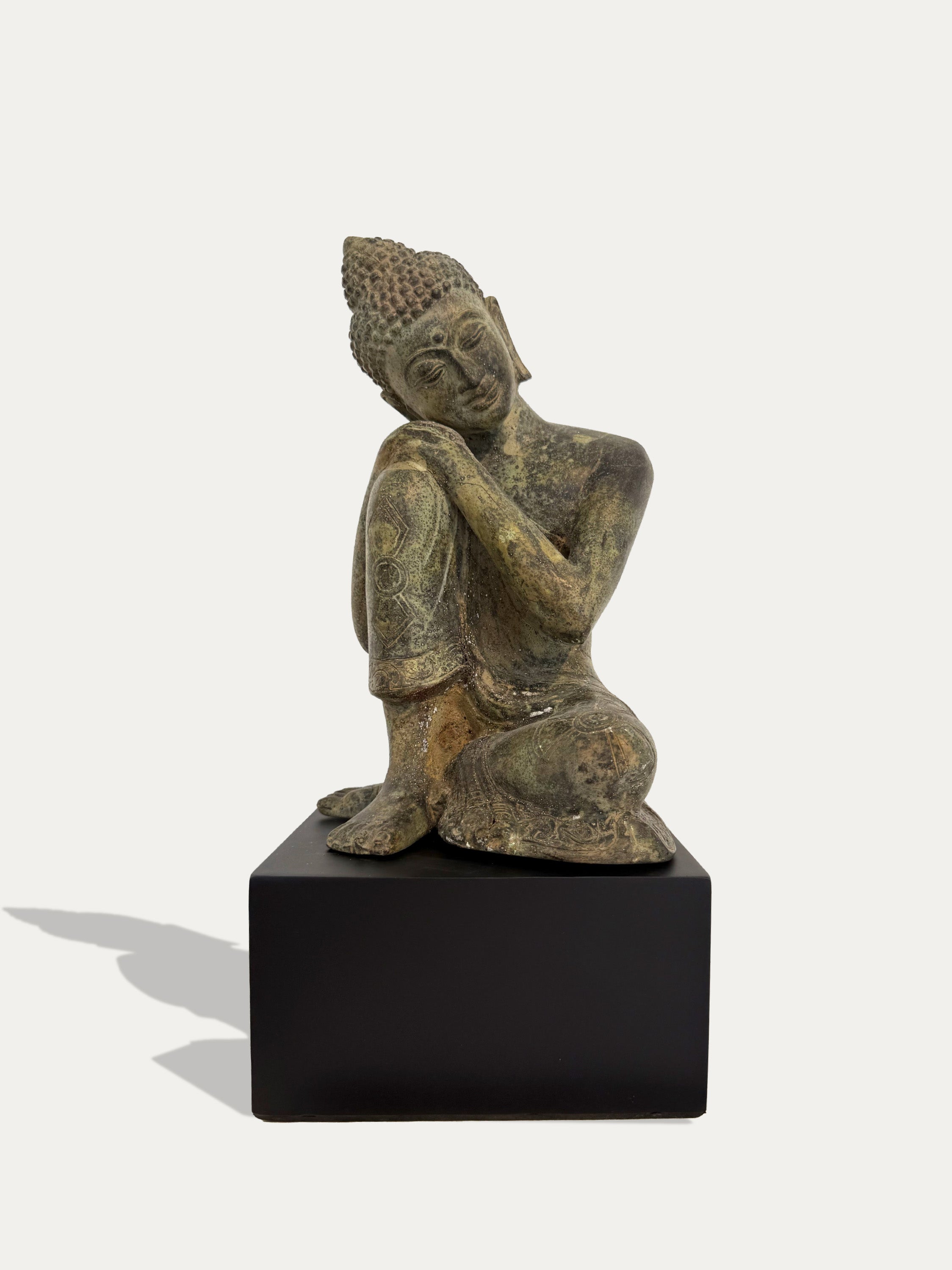 A bronze Buddha statue in a seated pose, displayed on a black rectangular pedestal against a light off-white background.