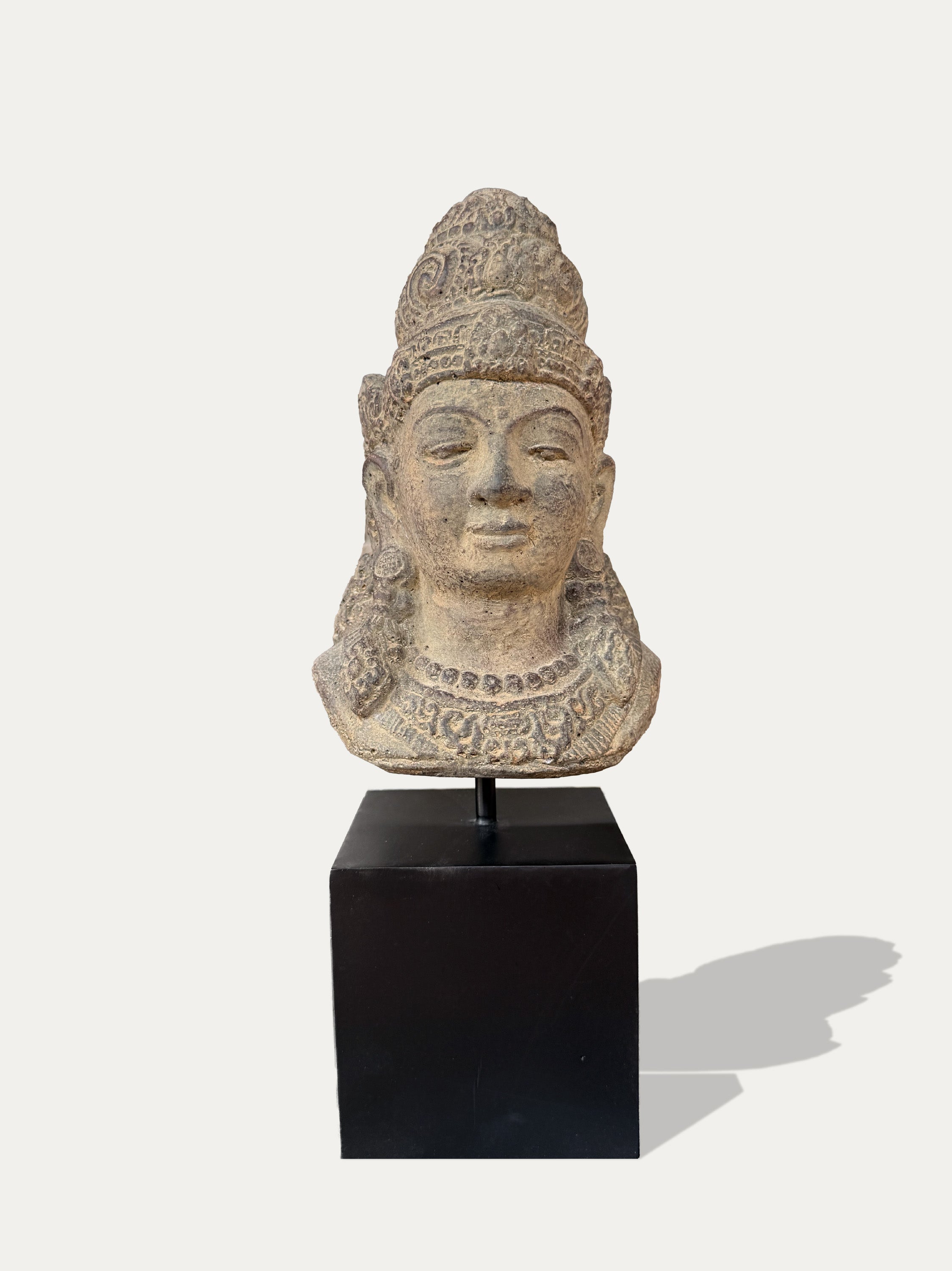 At stone Dewi Tara head sculpture with a detailed crown and necklace, mounted on a black display pedestal against a light gray background. 