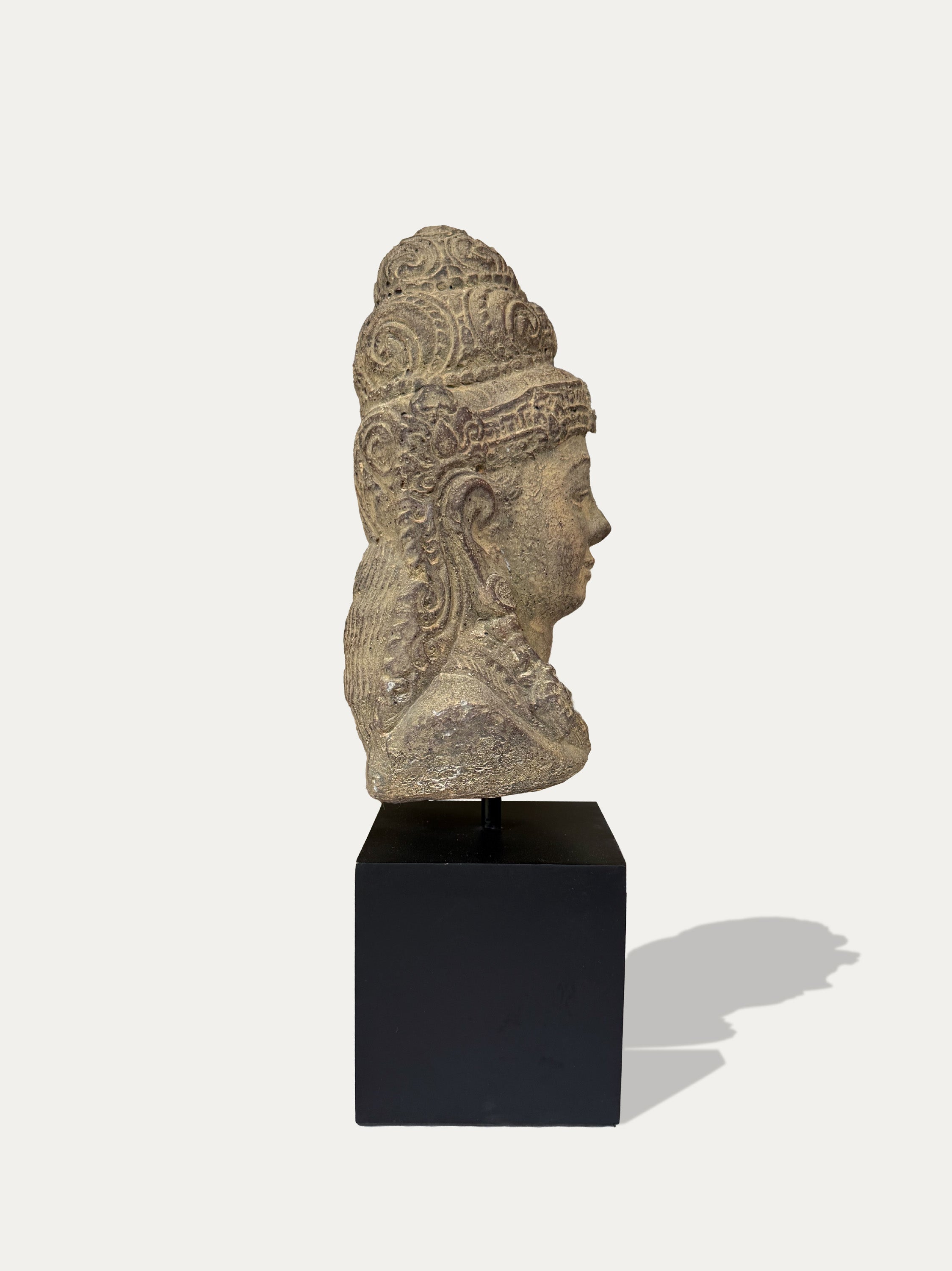 A stone Dewi Tara head sculpture in profile, mounted on a black display pedestal against a light gray background.