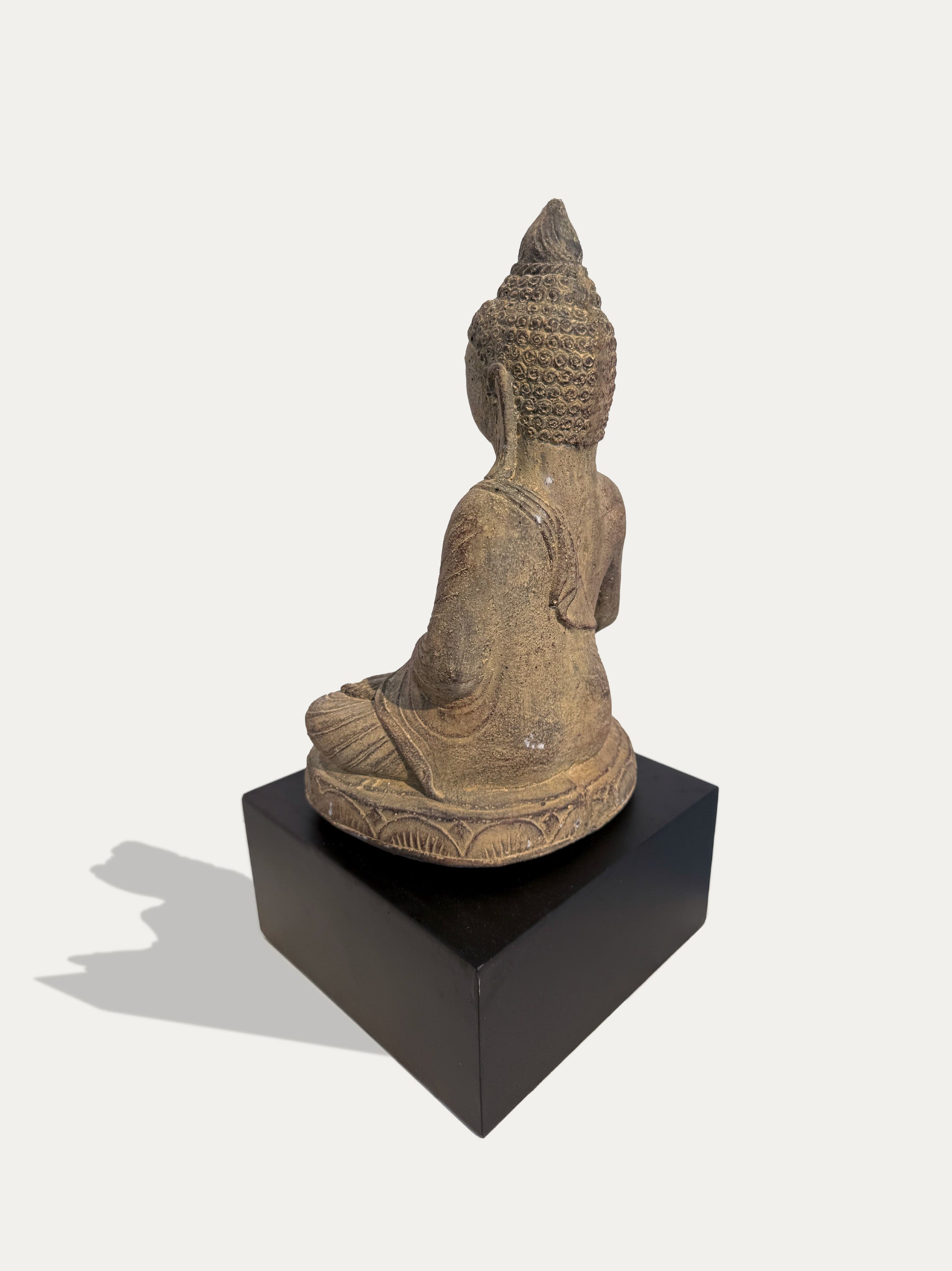 A seated Buddha statue in a weathered tan stone finish, shown from the back on a black rectangular pedestal against a light gray background.