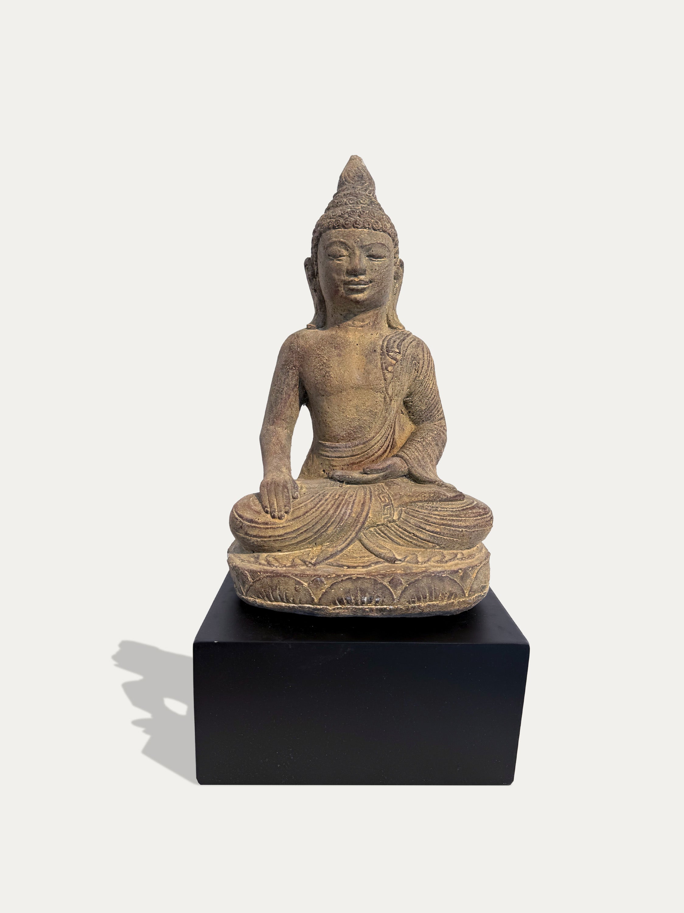 A seated Buddha statue in a weathered tan stone or terracotta finish, displayed on a solid black rectangular pedestal against a light gray background.