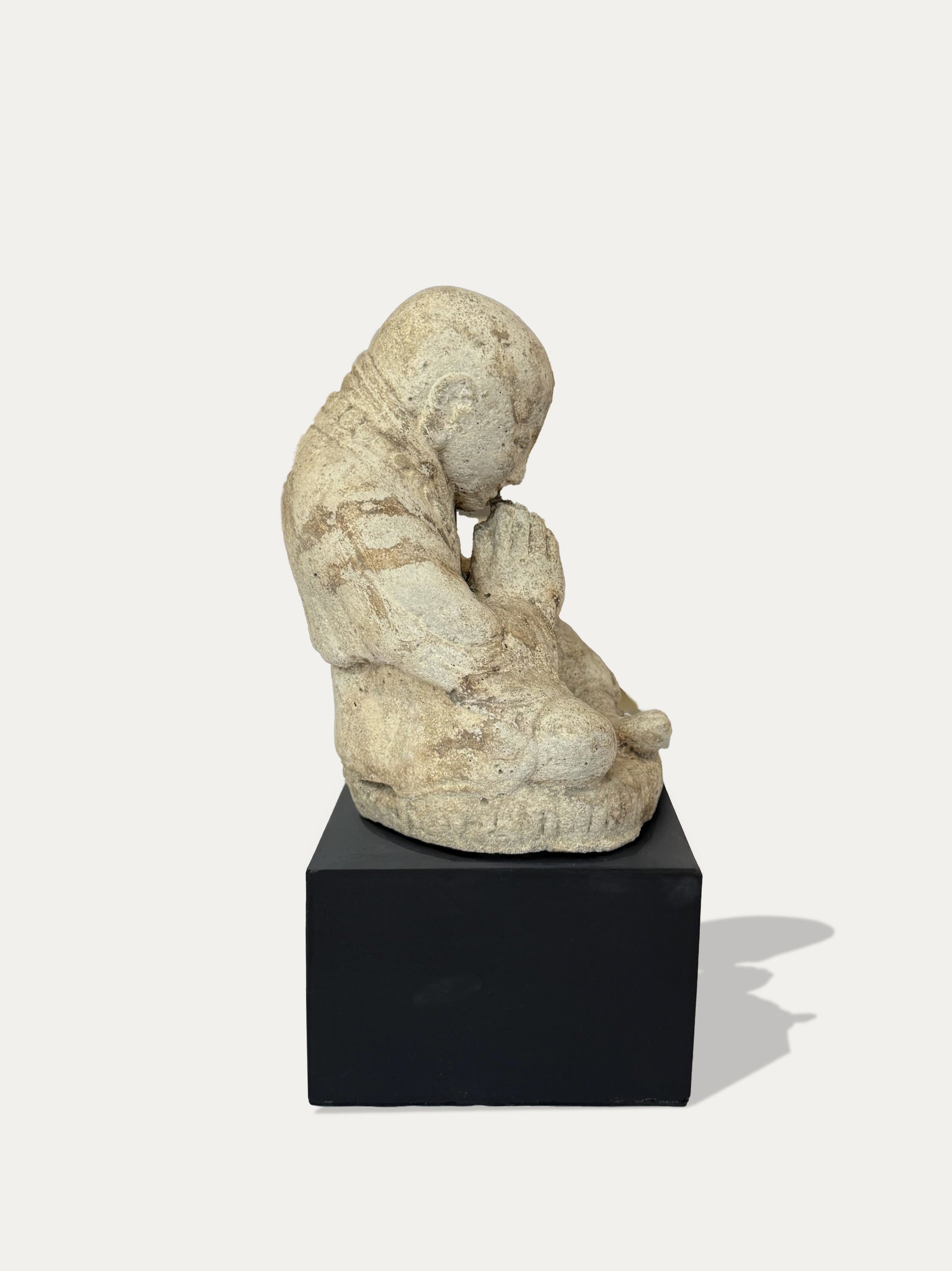 A small sculptural figurine of a seated child-like figure in a light stone or plaster finish, displayed on a black rectangular pedestal against a plain light background.
