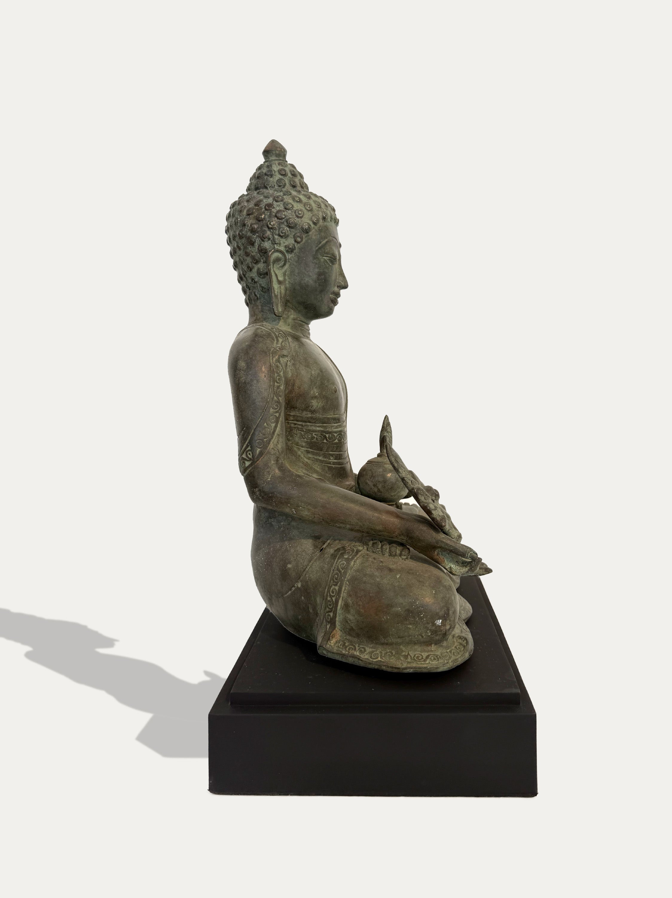 A seated Buddha statue in an aged bronze finish, positioned in profile on a black rectangular pedestal against a light gray background.