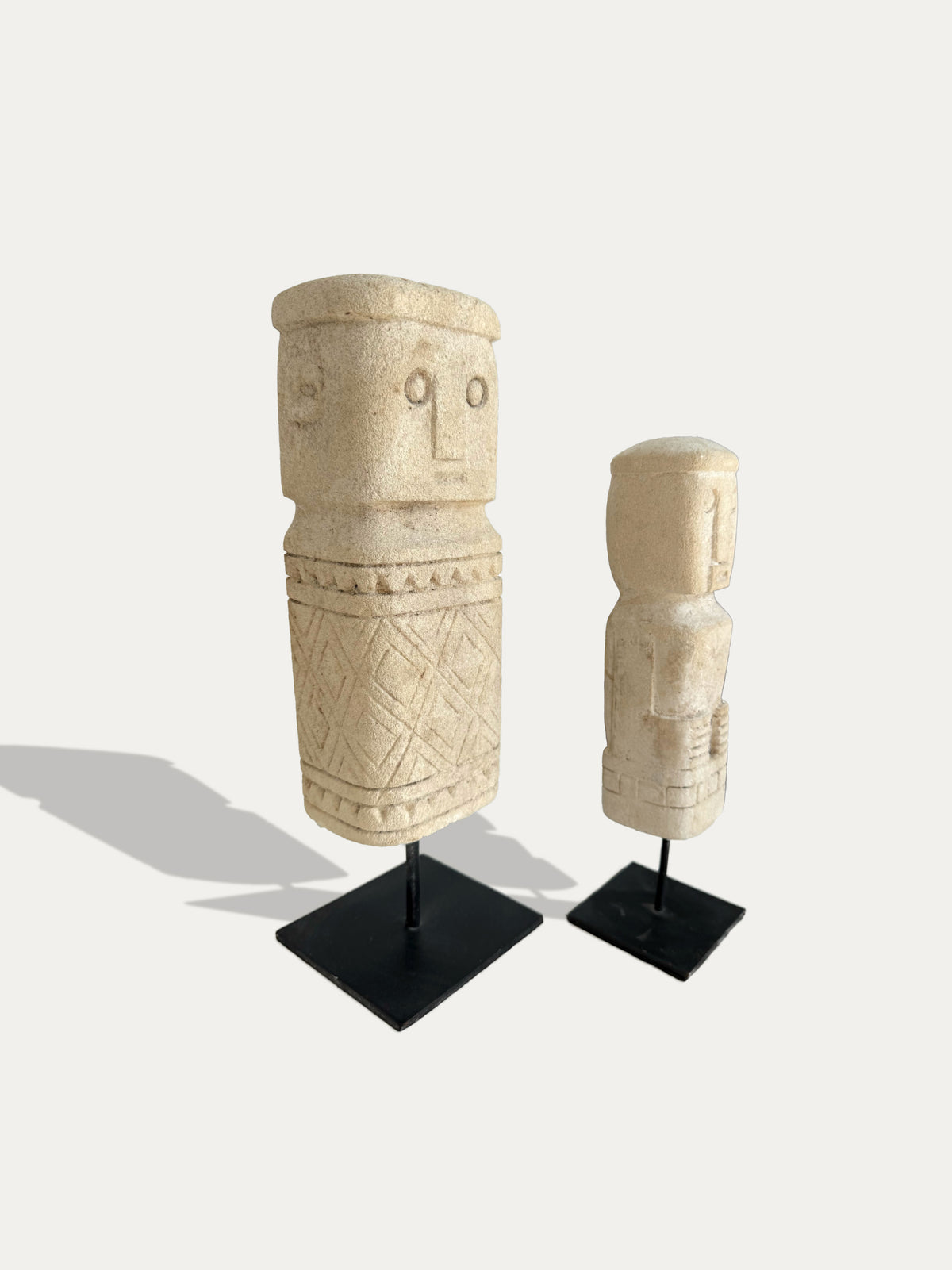 Angled view of two Timor limestone statues showing carved faces and geometric patterns on black metal stands.