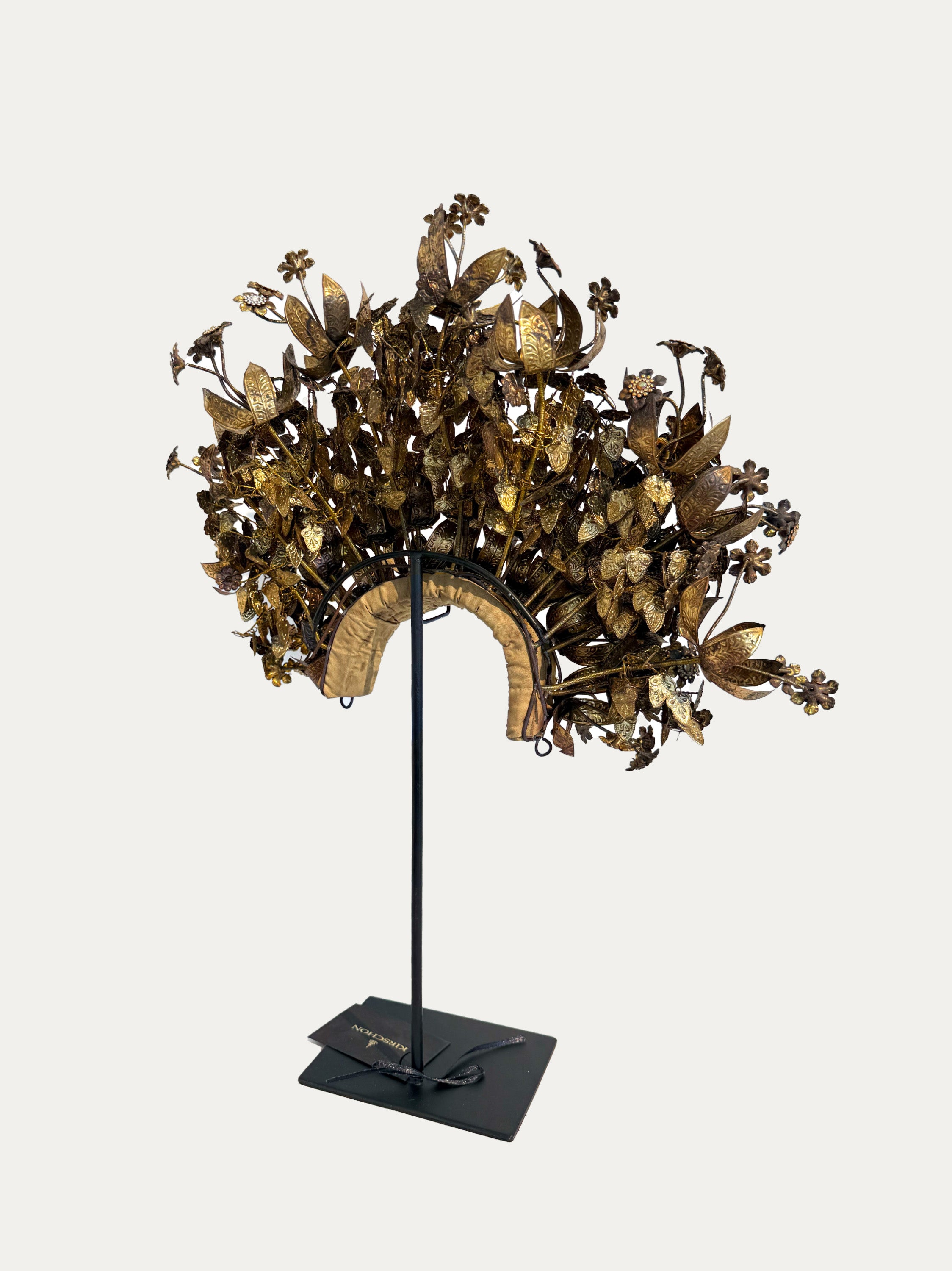 An ornate gold-toned floral metal headpiece displayed on a black stand against a light gray background.