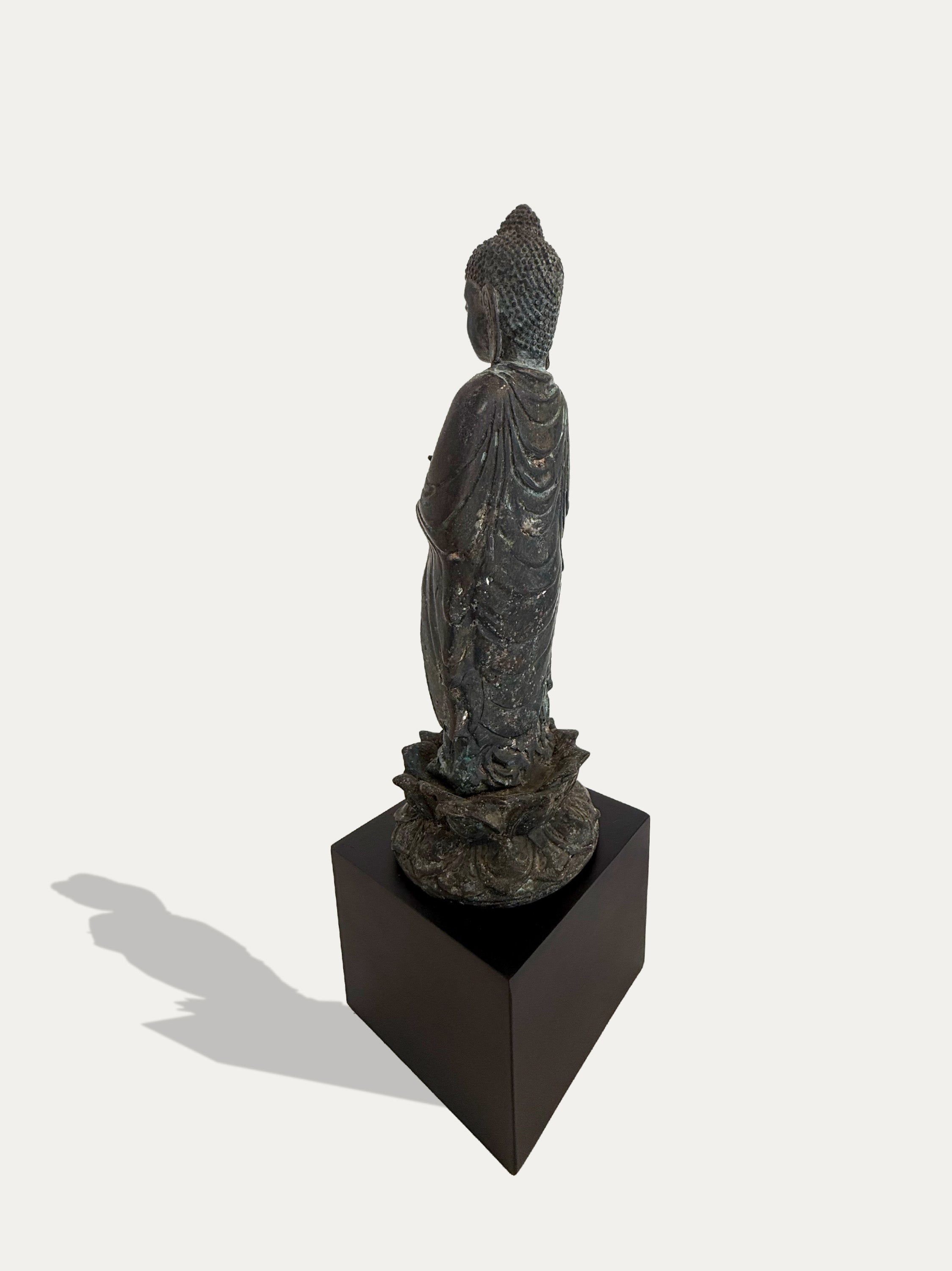 A dark metallic Buddha statue shown from the back, mounted on a black rectangular pedestal against a light gray background.