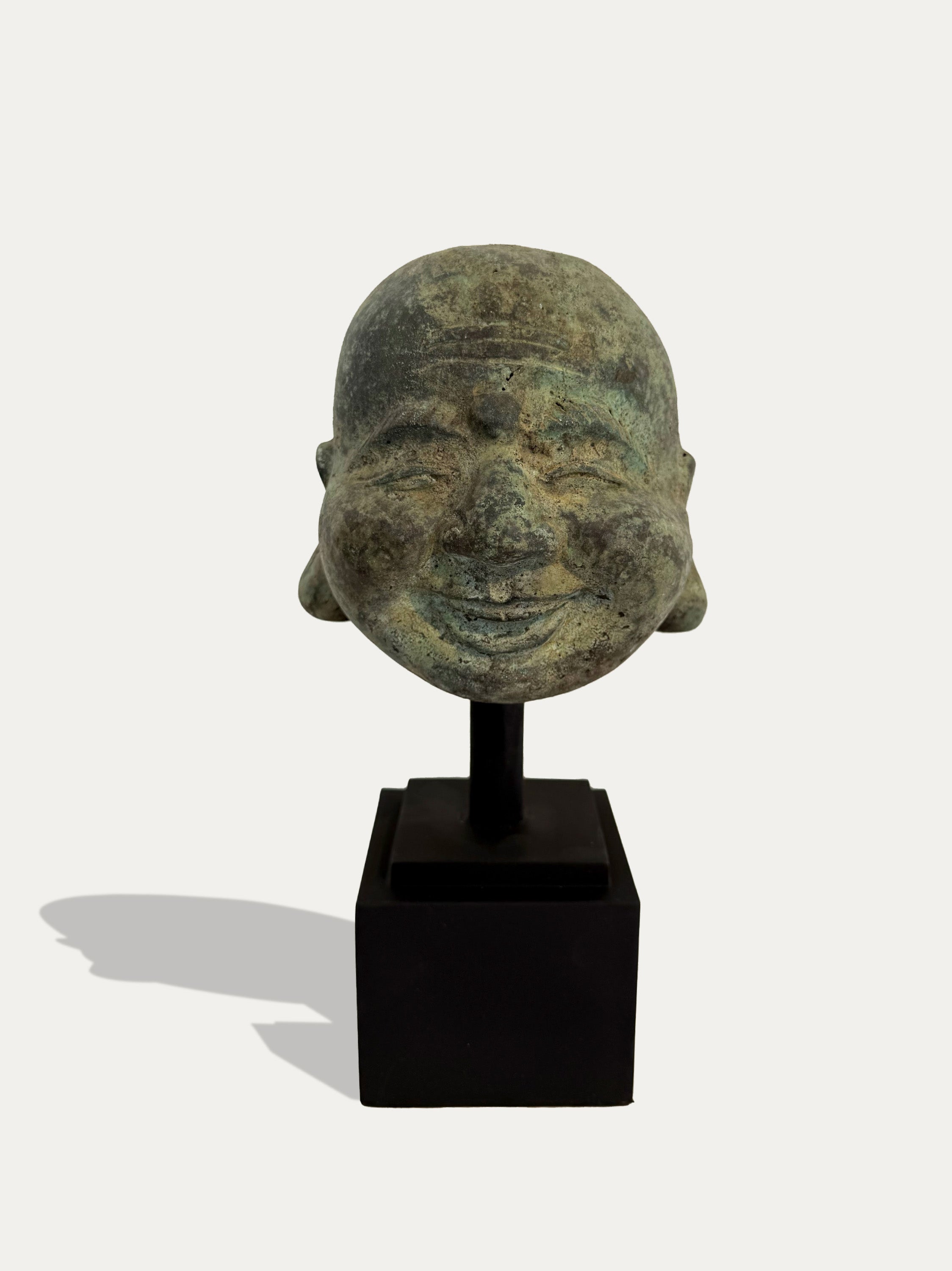 An ancient-style bronze Buddha head sculpture with a green patina, mounted on a black display stand against a light gray background.