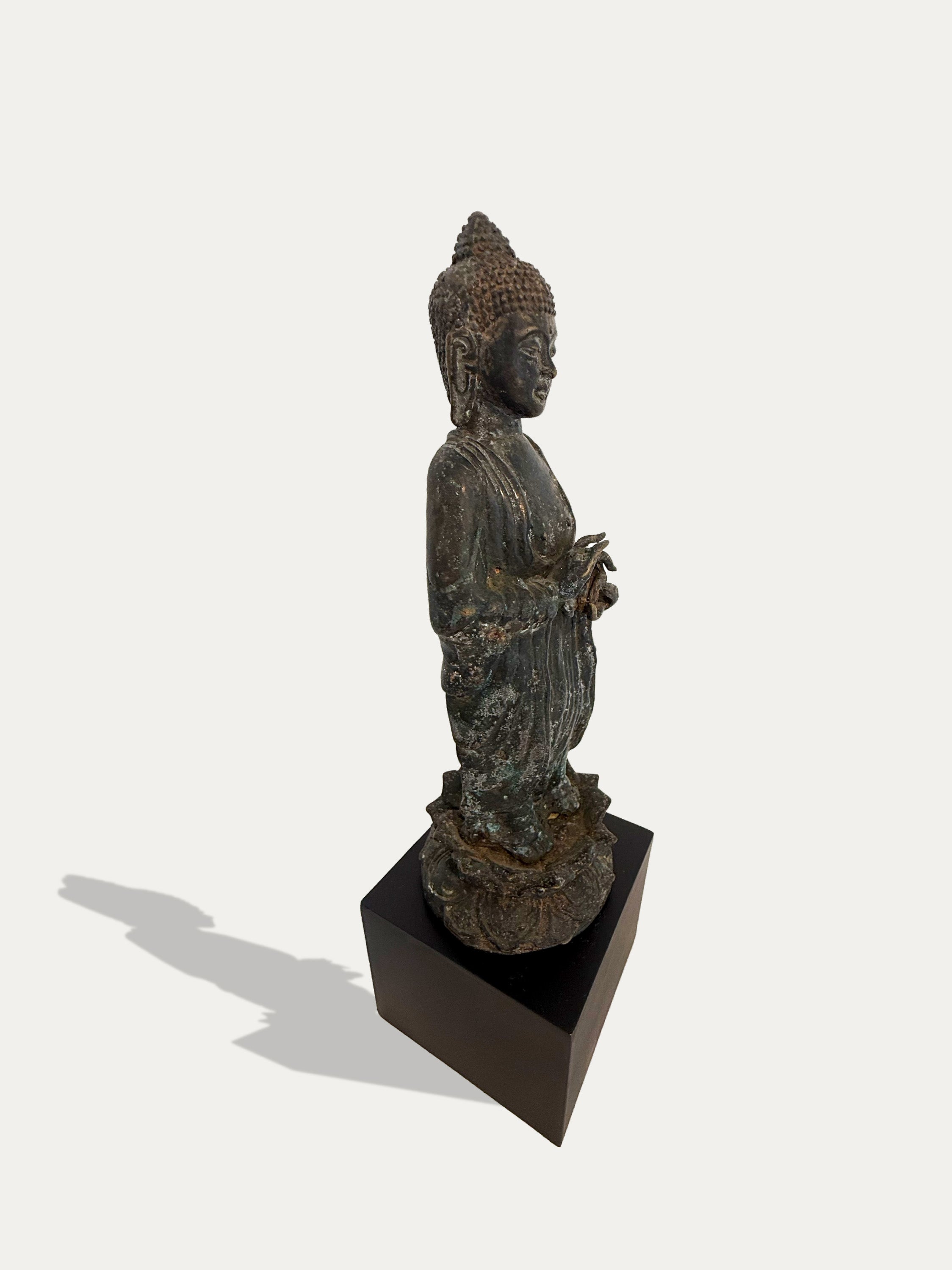 An antique-style bronze Buddha statue standing on a dark square pedestal, photographed against a plain light background with a soft shadow cast to the left.