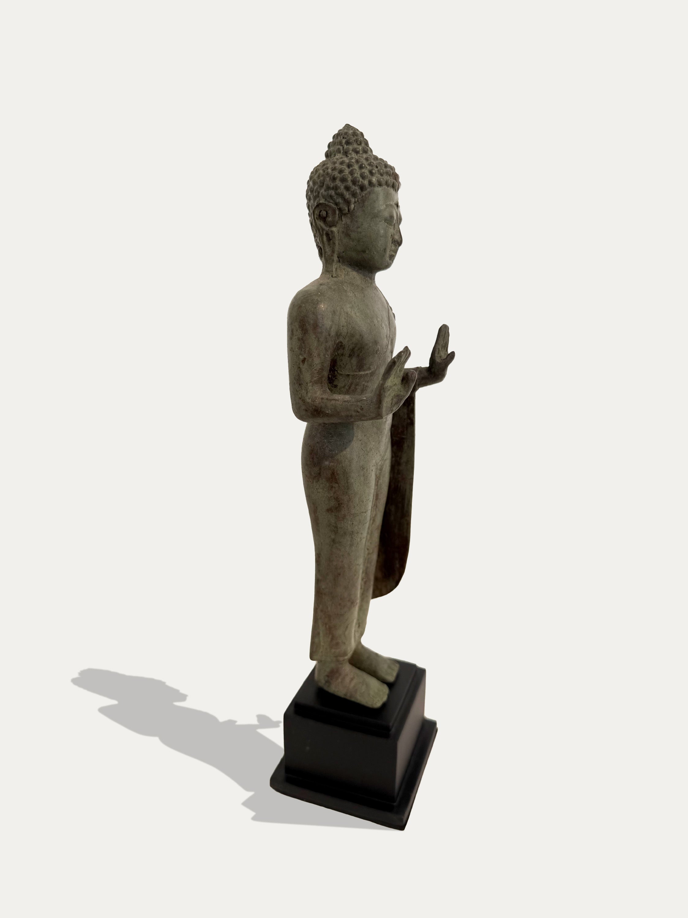 A standing Buddha statue in a weathered gray-green stone finish, mounted on a black rectangular base against a light gray background with a soft shadow.